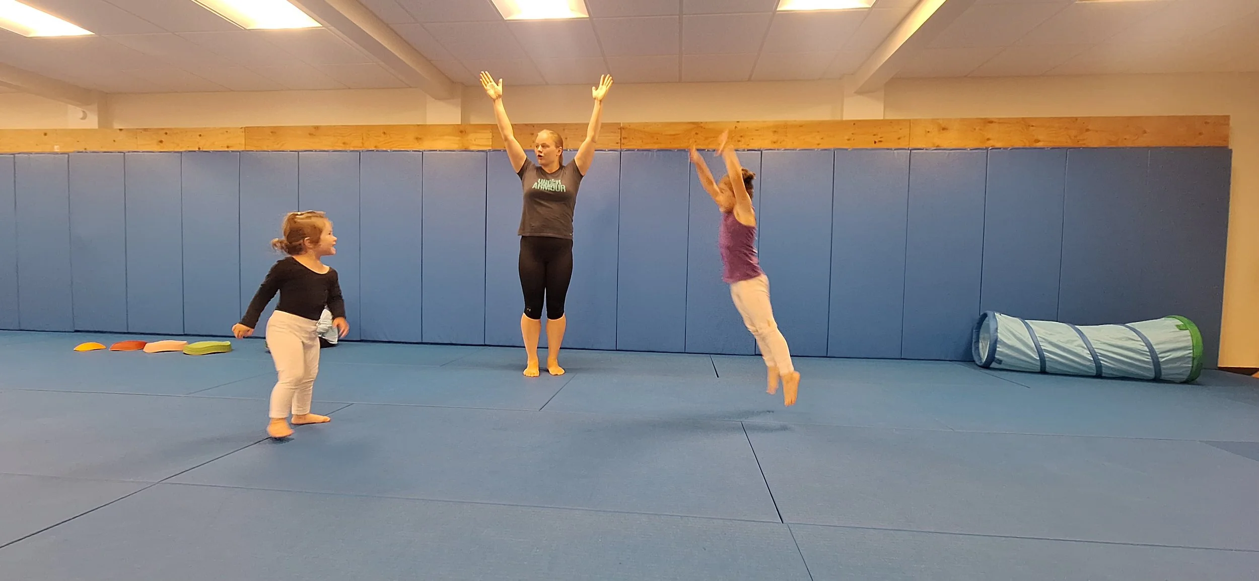 A woman practicing aerial acrobatics with two young girls in a gym. The woman is mid-air, holding a hammock or trapeze, while the girls watch and smile. The gym has blue padded walls and a blue floor.