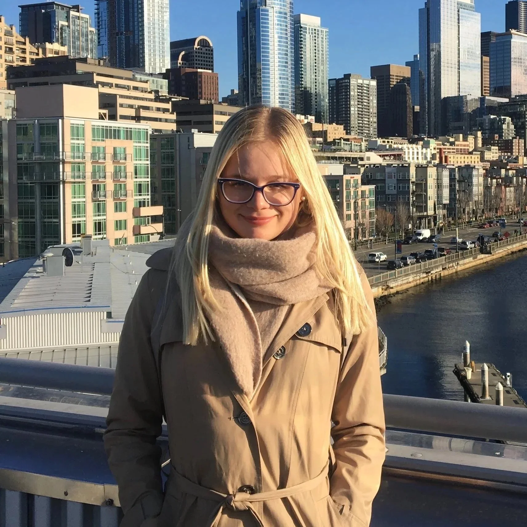A woman with blonde hair and glasses wearing a beige coat and scarf standing on a bridge in front of a cityscape with modern high-rise buildings and a river.