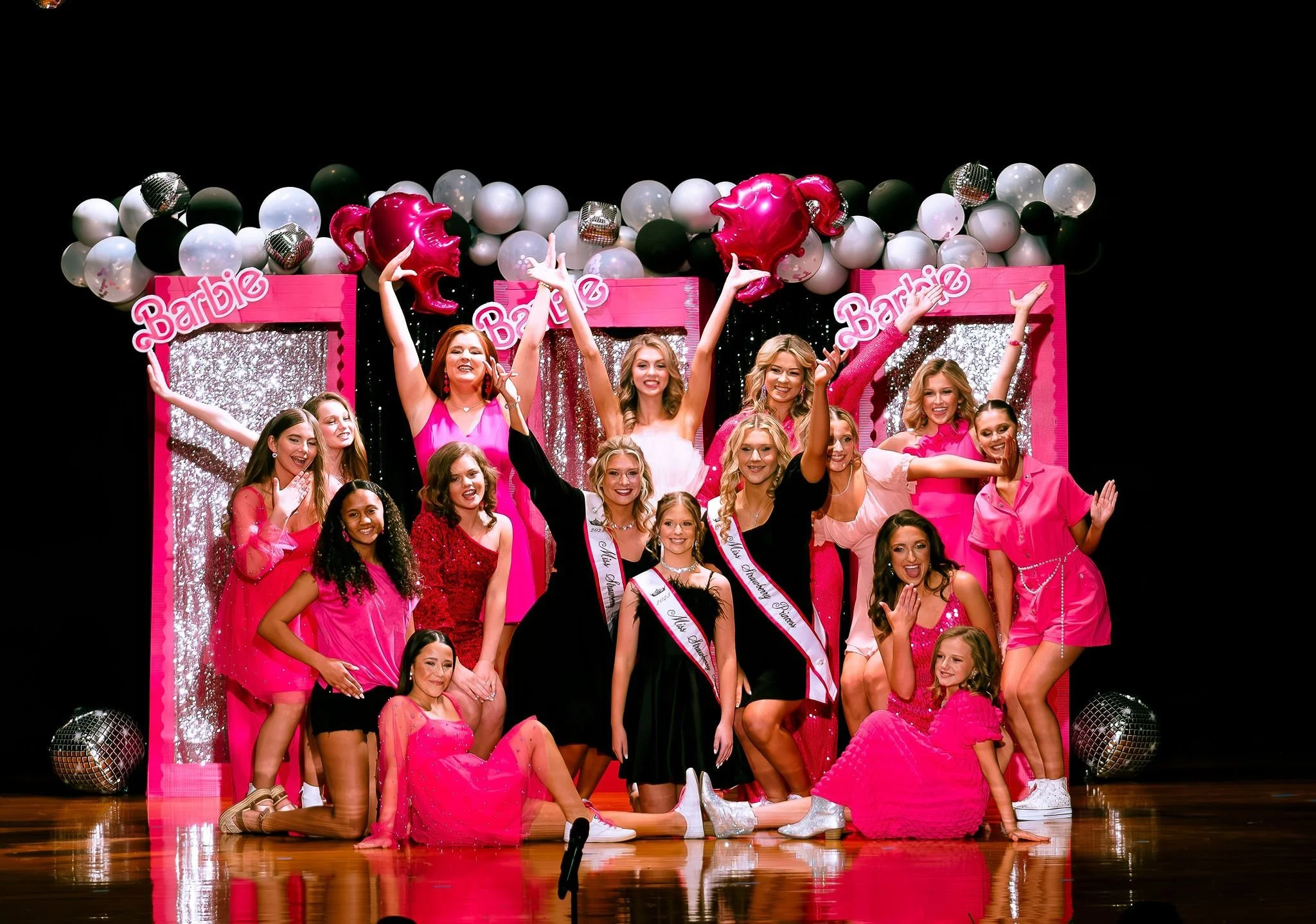 Group of young women and girls celebrating on stage at a Barbie-themed event, with pink decorations, balloons, and sashes. Most are dressed in pink or black outfits, smiling and posing.