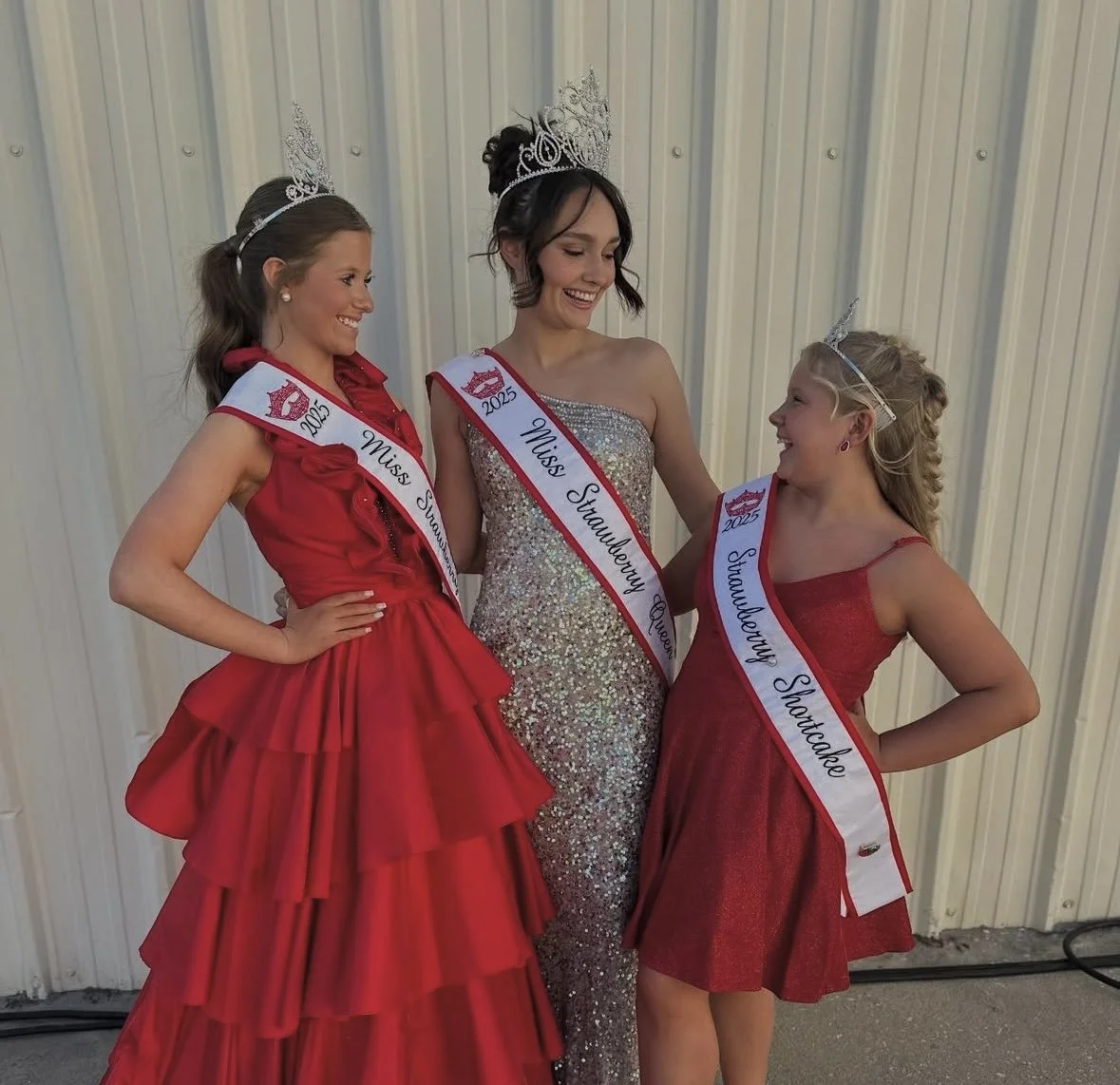Three young women dressed in formal gowns wearing sashes and tiaras, celebrating a pageant event. The woman in the middle is Miss Strawberry Queen, the woman on the left is Miss Strawberry, and the woman on the right is Shandewy Shortcake, all from 2