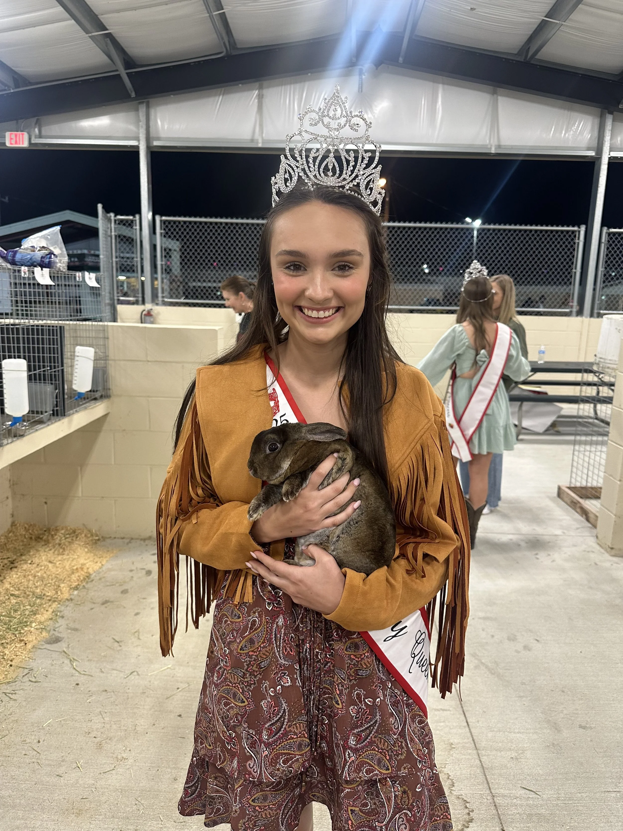 A young woman wearing a crown and a sash, dressed in Western-style clothing, is holding a brown rabbit at an indoor event. She is smiling at the camera, and in the background, other women can be seen, some of whom are also wearing sashes.