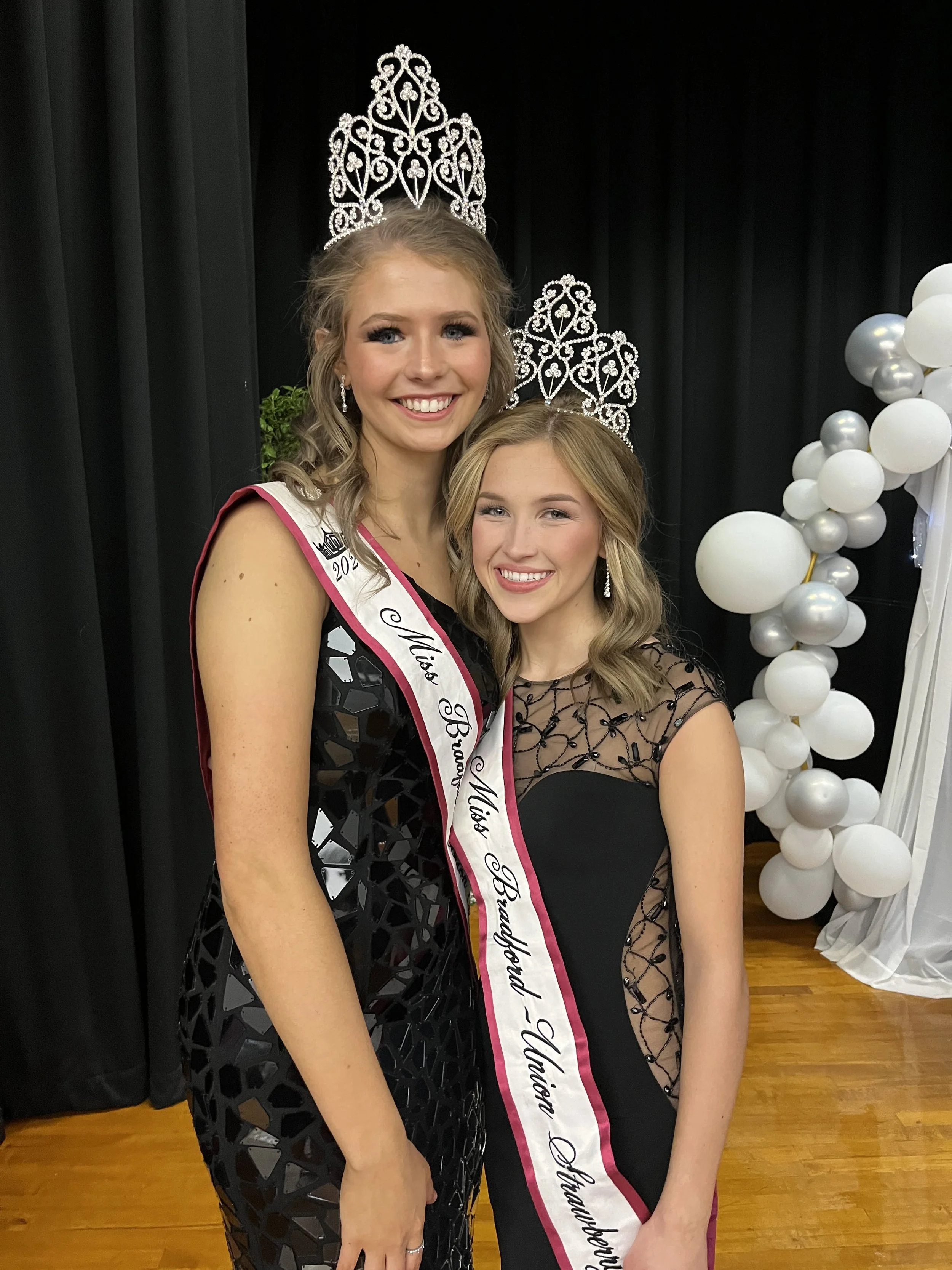 Two young women dressed in black evening gowns and wearing tiaras, with one wearing a sash that reads 'Miss Brunswick - North Brunswick, Miss Badland - North Brunswick.' They are smiling and standing in front of black curtains and white balloon decor
