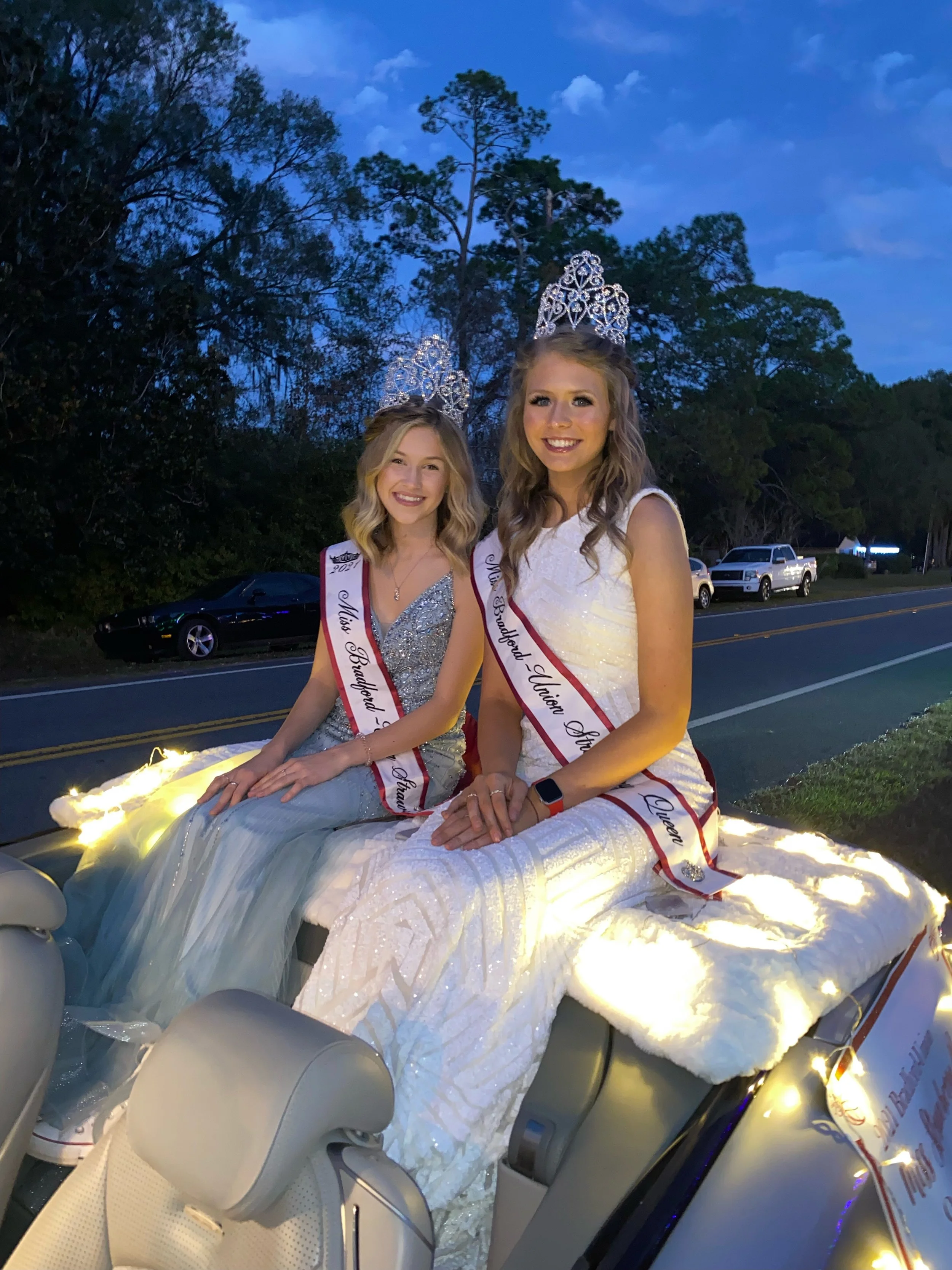 Two young women wearing tiaras and sashes sitting on a decorated vehicle during a parade, with a background of trees and a few parked cars in the evening.
