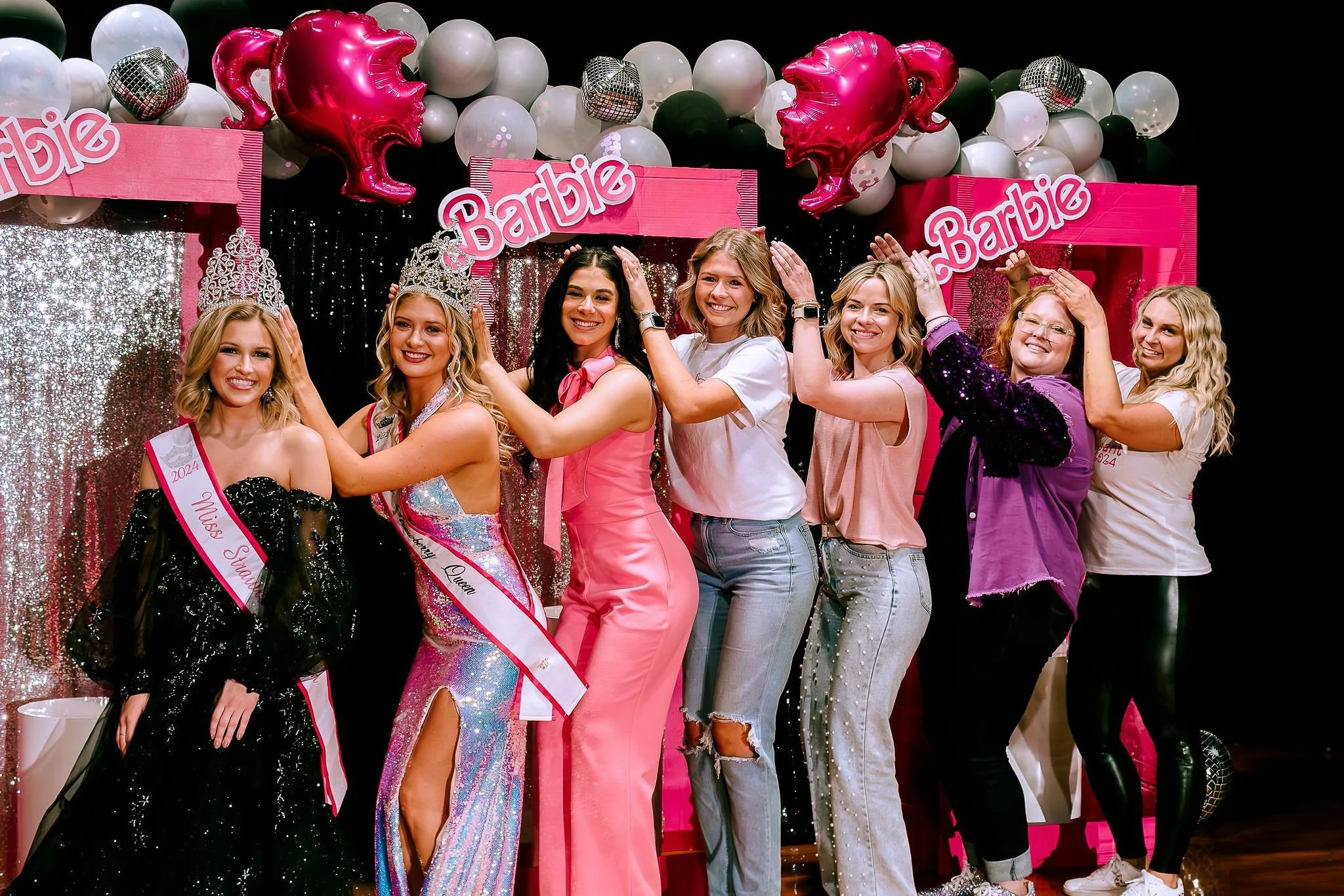 A group of women at a beauty pageant celebration, with two contestants wearing crowns and sashes, surrounded by pink and white balloons and a glittery backdrop.