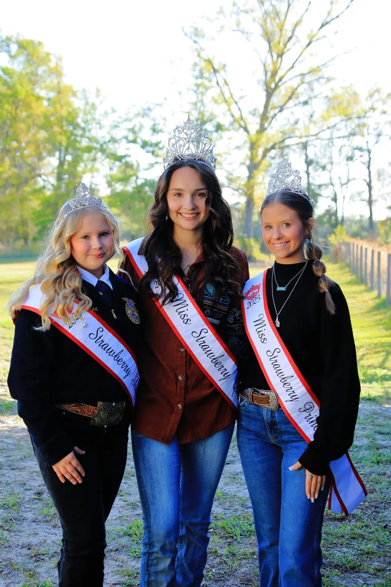 Three young women wearing crowns and sashes posing outdoors, celebrating a strawberry festival in 2025.
