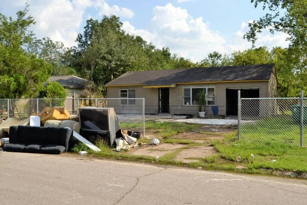 A house with a broken driveway and trash, furniture, and debris outside, surrounded by chain-link fences.