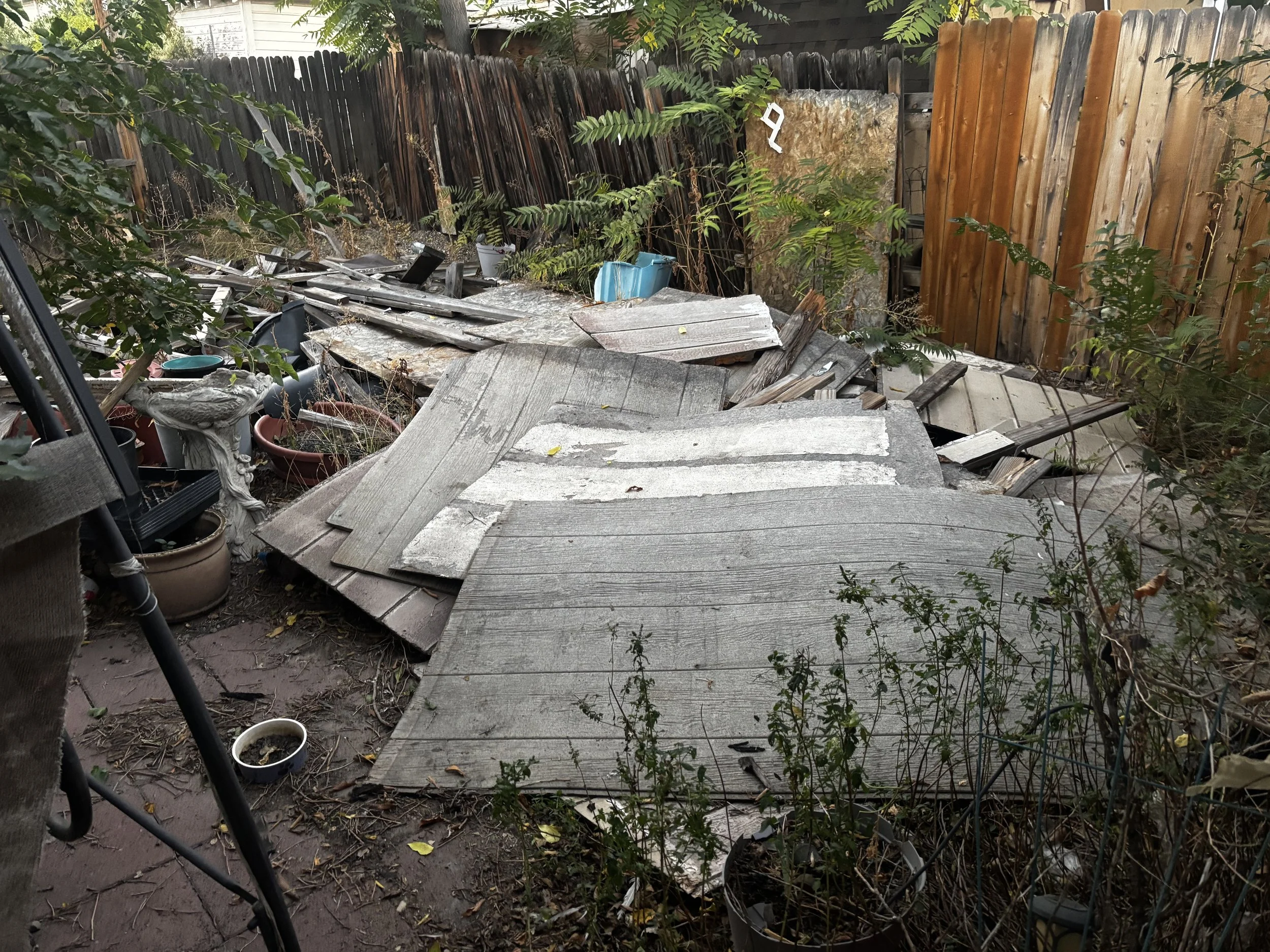A backyard with a collapsed wooden deck or shed, surrounded by planters and overgrown plants, enclosed by a wooden fence.