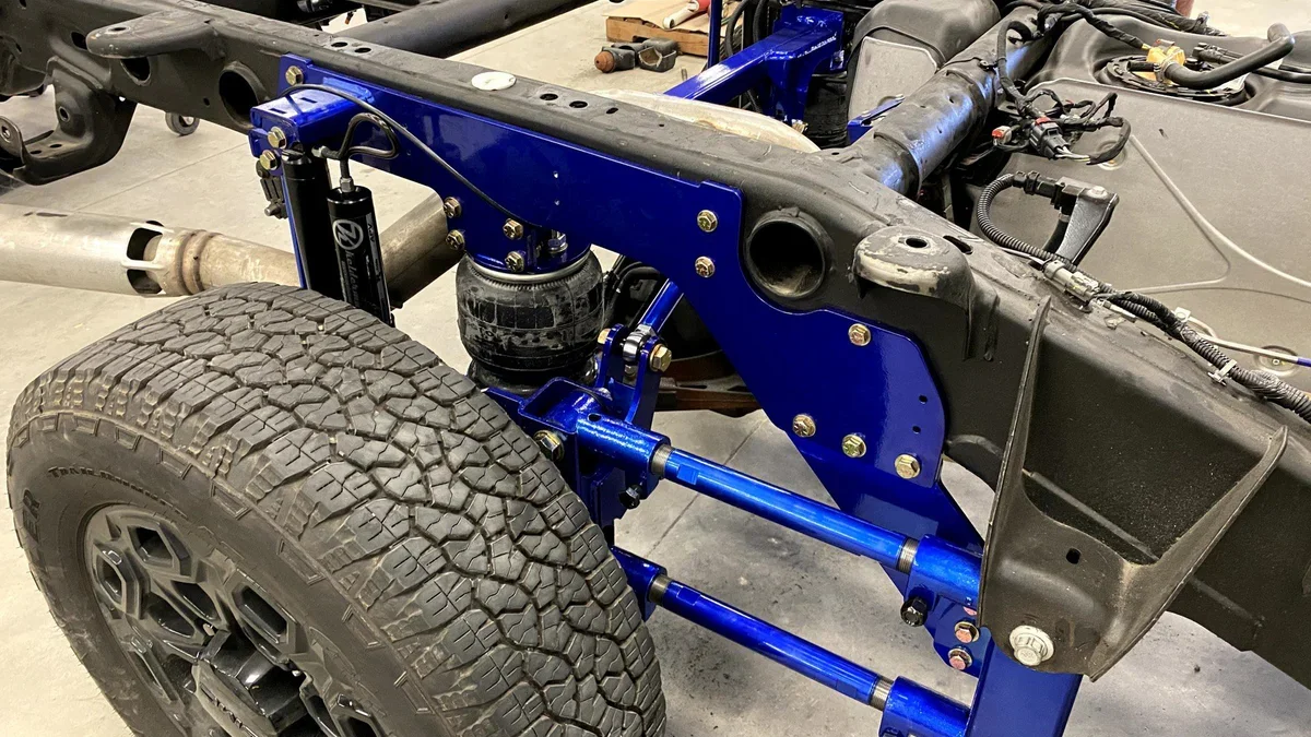 Close-up of a vehicle chassis showing suspension components, blue-painted suspension arms, a black shock absorber, large off-road tire, and part of the engine bay.