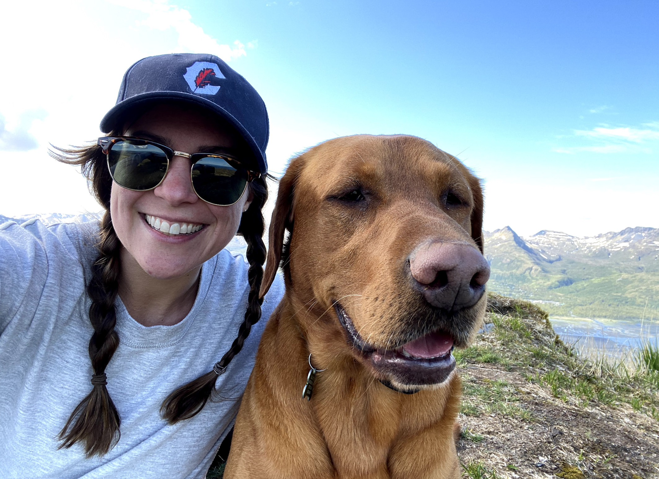 A woman wearing sunglasses and a baseball cap smiling next to a large brown dog, with mountains and blue sky in the background.