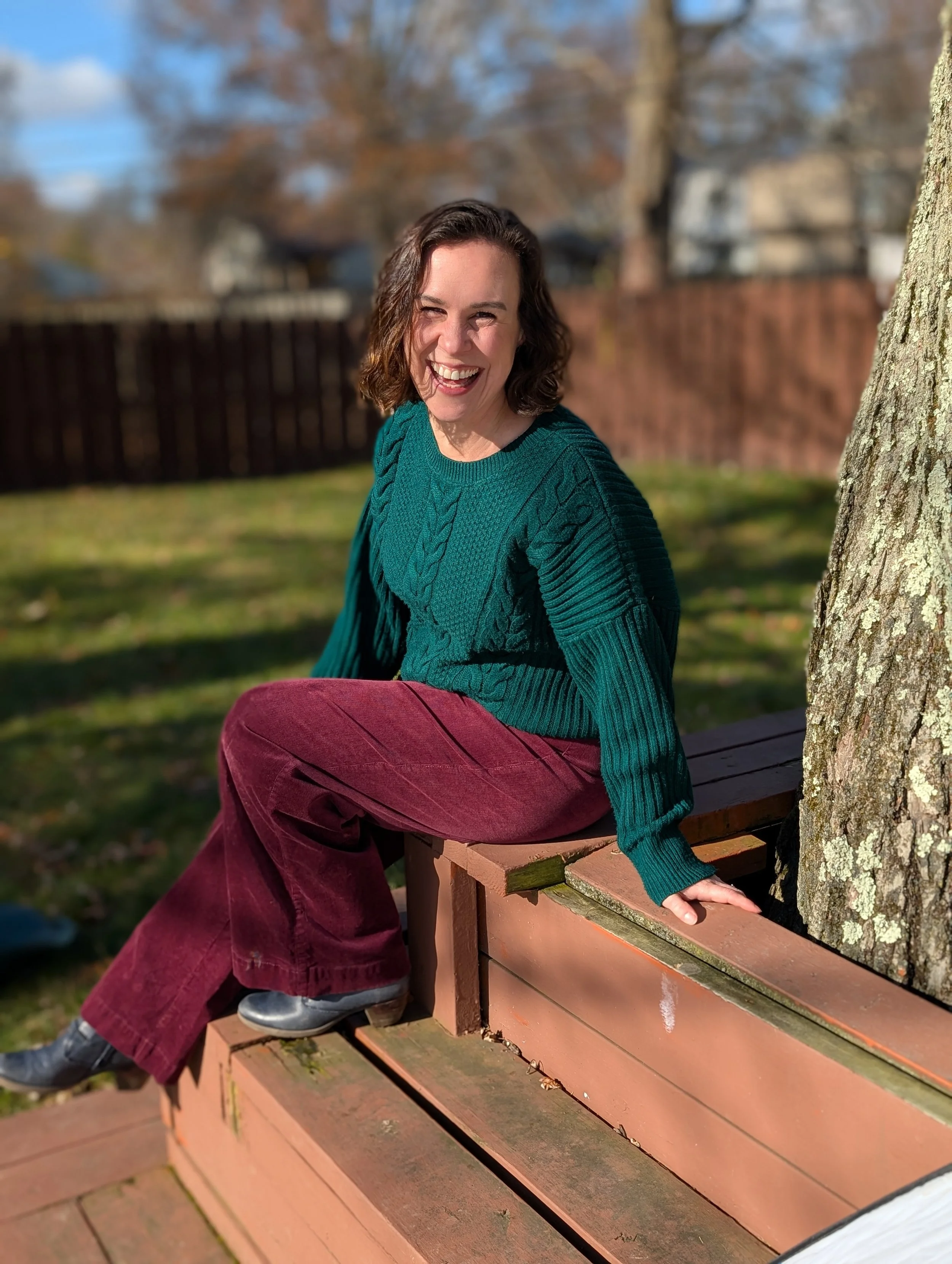A woman sitting on a wooden bench outdoors, smiling and wearing a teal sweater and maroon pants. She is positioned near a tree and a backyard fence, with a blurred background of trees and houses on a sunny day.