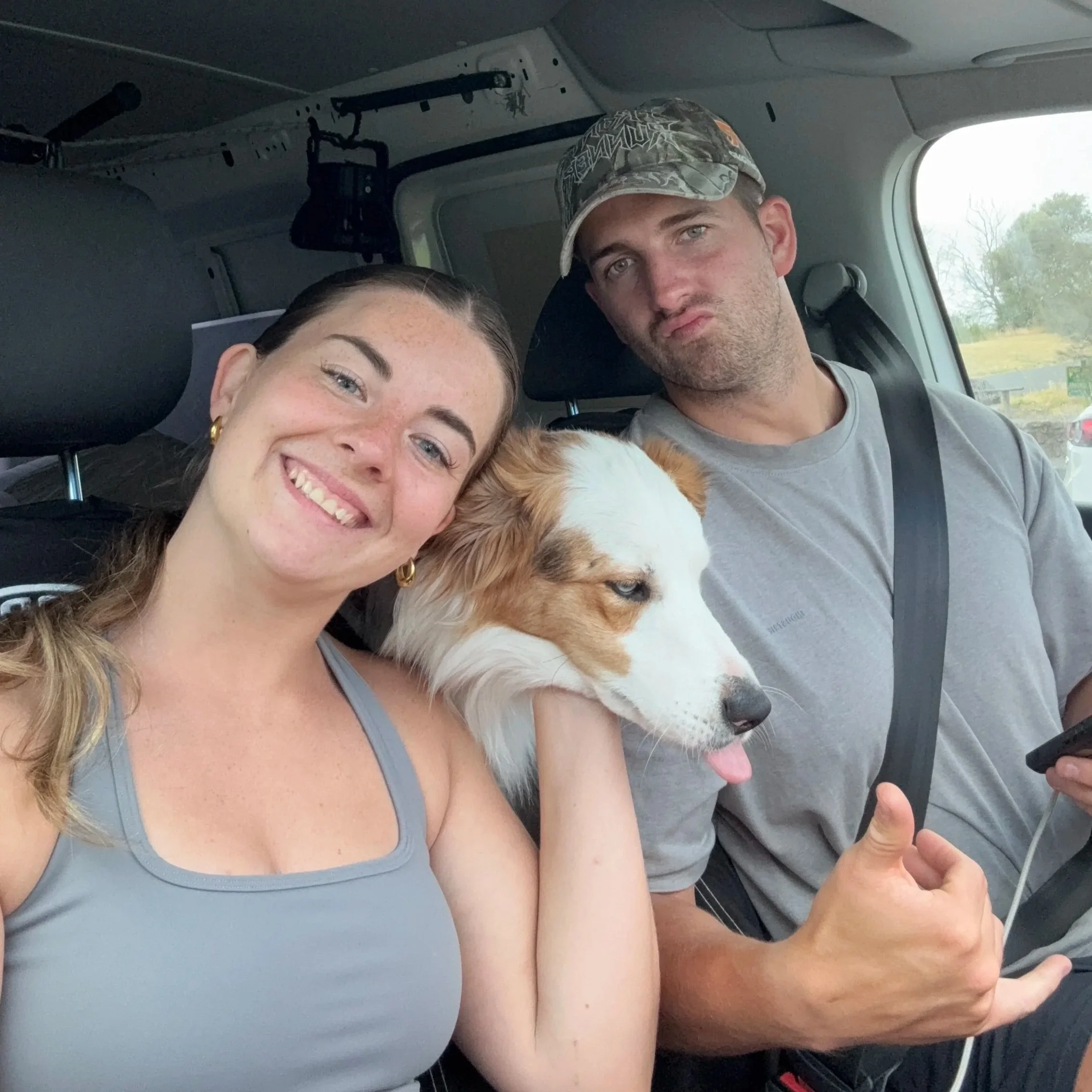 A selfie of a young woman and a young man inside a vehicle, with a dog between them. The woman is smiling, and the man is looking at the camera with a serious expression. The woman has long, dark hair and is wearing a gray tank top. The man has short hair, facial hair, and is wearing a gray shirt and a camouflage hat. The dog has white and brown fur and is partially visible.