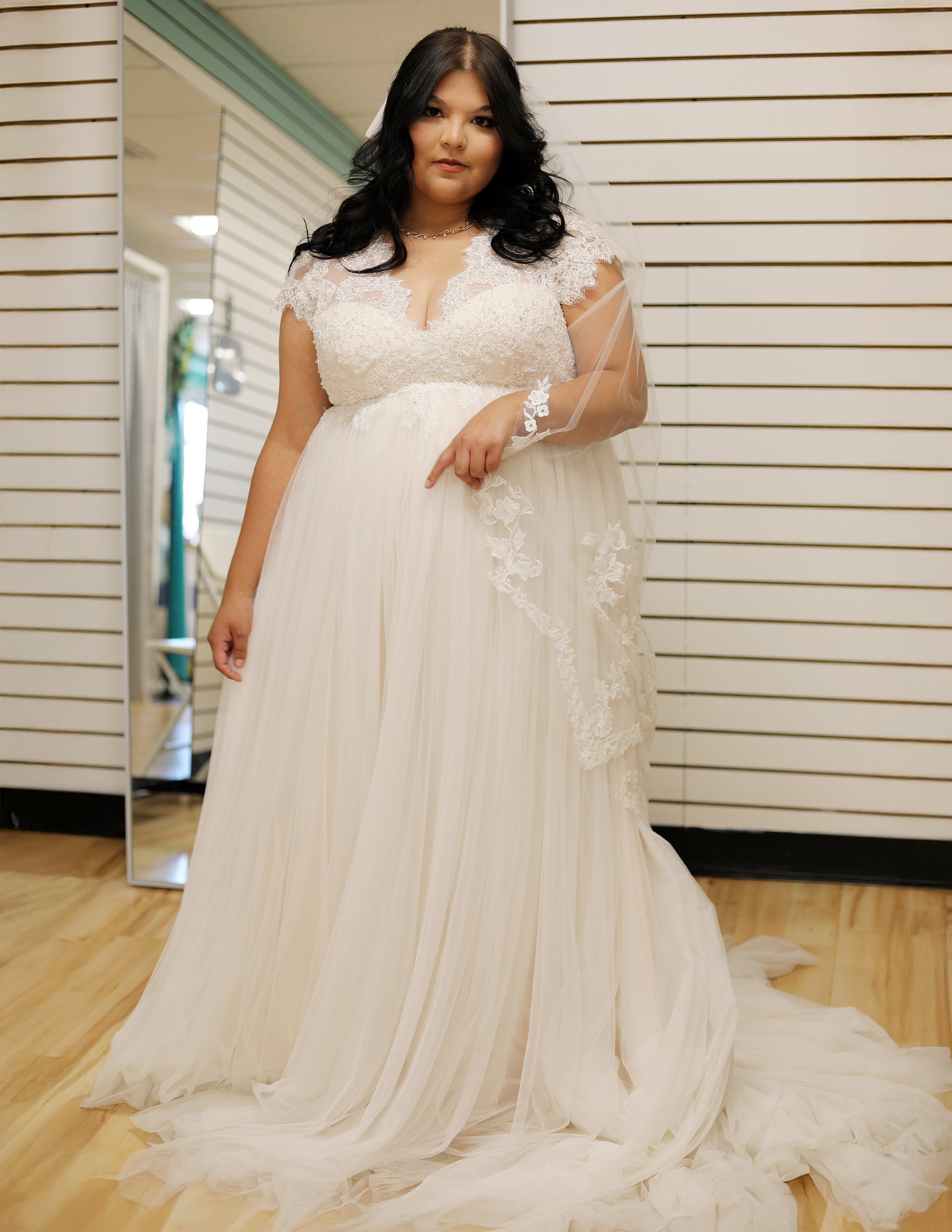 A woman with long dark hair wearing a wedding dress with lace and tulle, standing indoors against a wall with horizontal wooden slats.