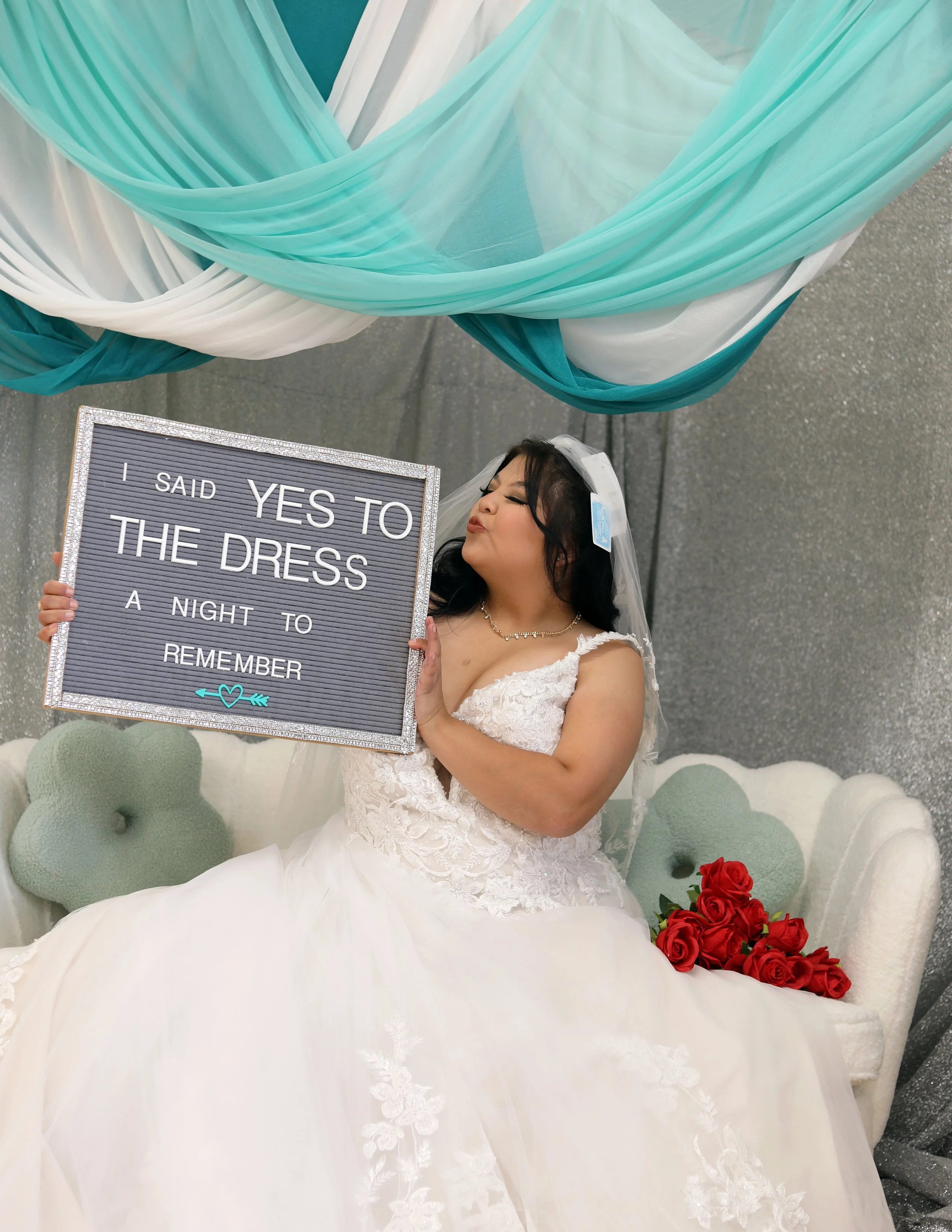 A woman in a wedding dress holding a sign that reads 'I said YES to the dress a night to remember.' She is sitting on a white couch with red roses beside her, and there are decorative drapes overhead.