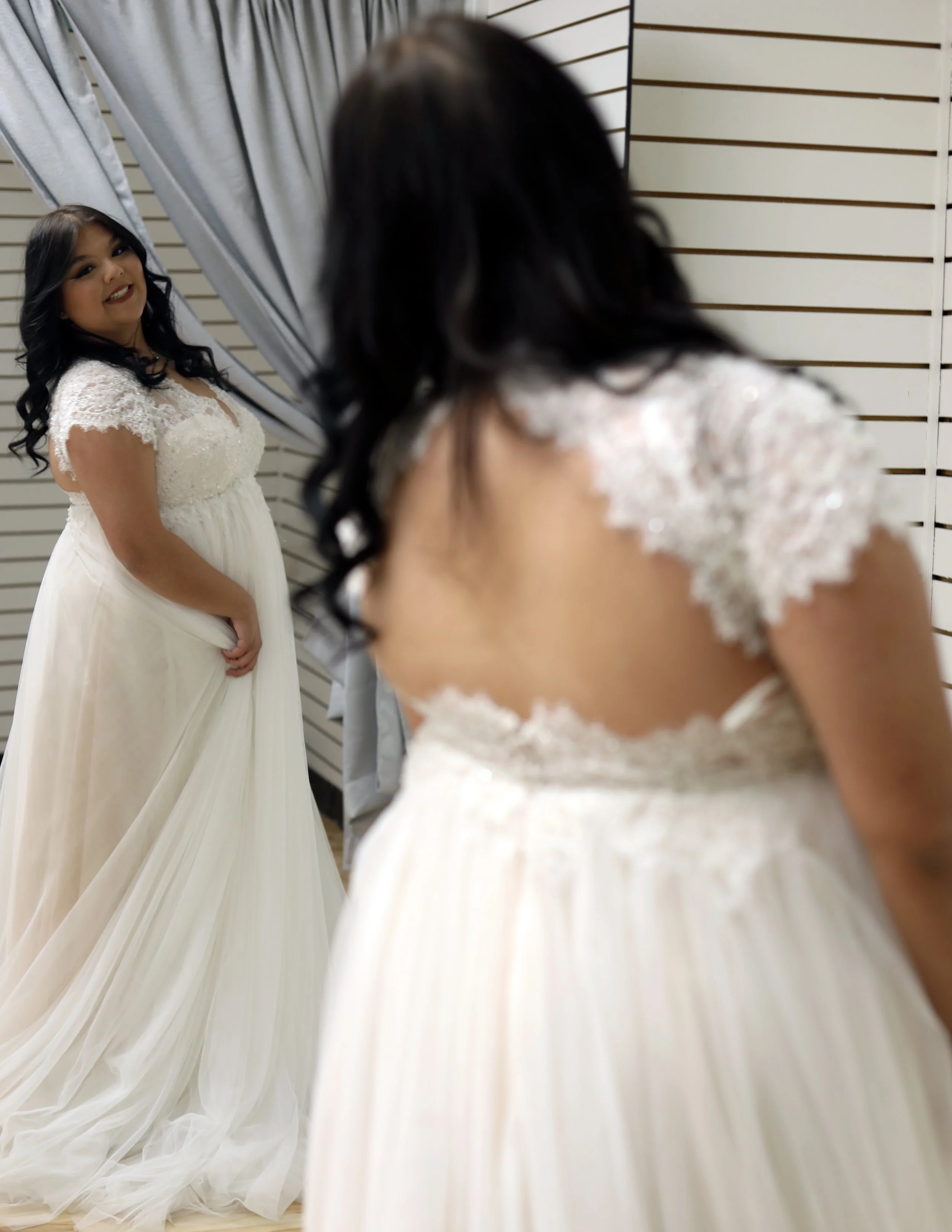 A woman in a white lace wedding dress looking at herself in a mirror with a smile, reflecting her image.