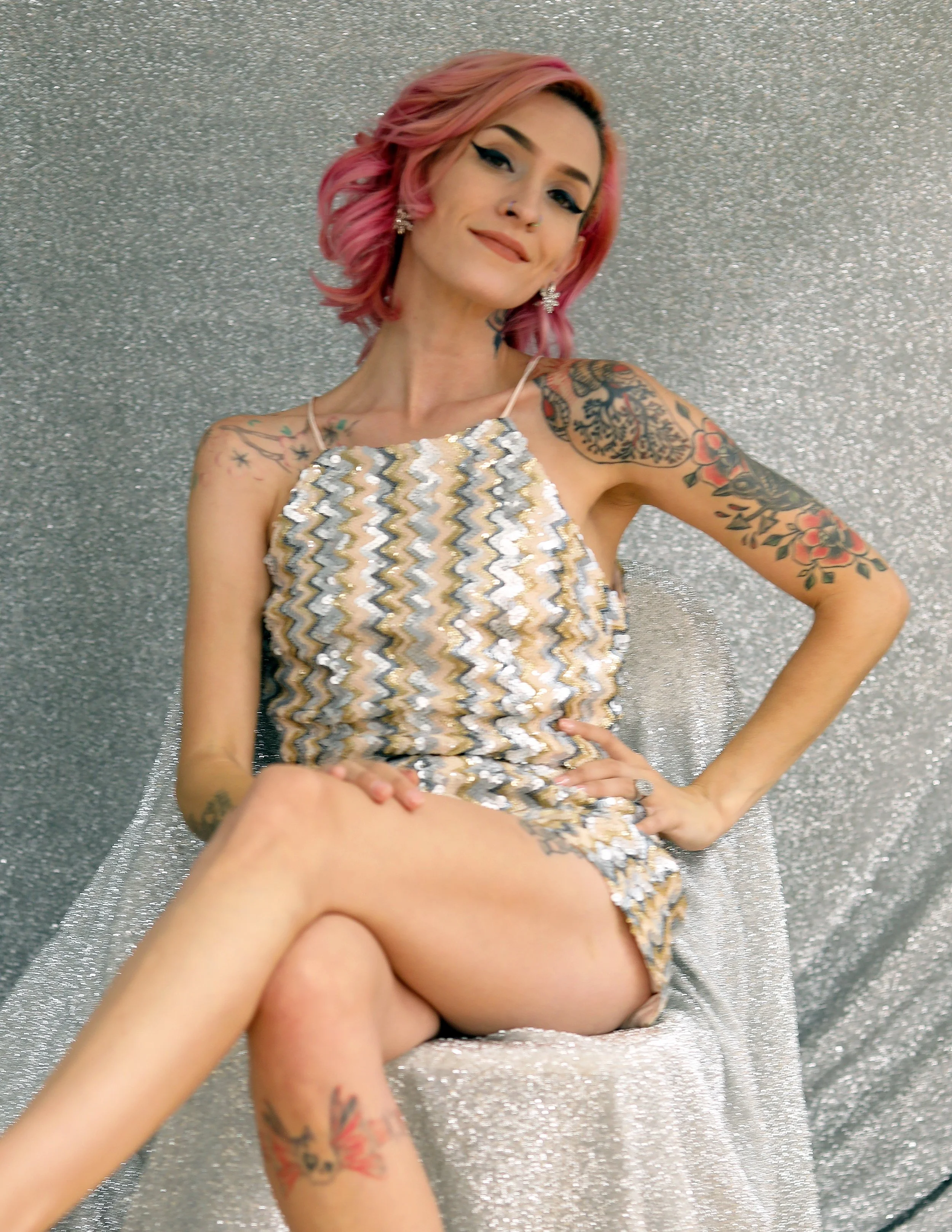 Woman with pink hair and tattoos sitting on a sparkly silver chair, wearing a patterned dress and smiling.