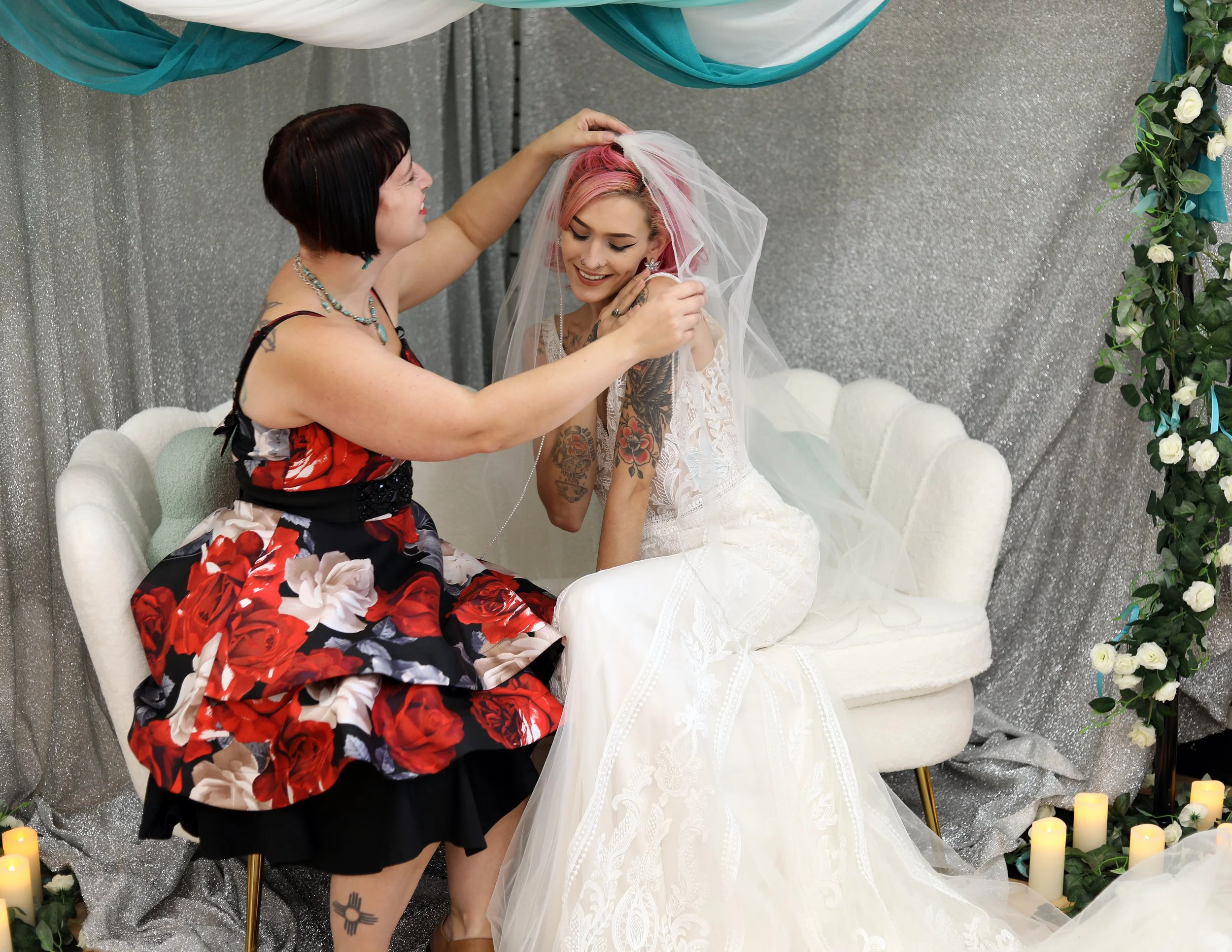 A bride with pink hair sitting on a white velvet couch, smiling as an older woman helps her with her veil. The background is decorated with silver fabric, green leaves, white flowers, and lit candles on the floor.