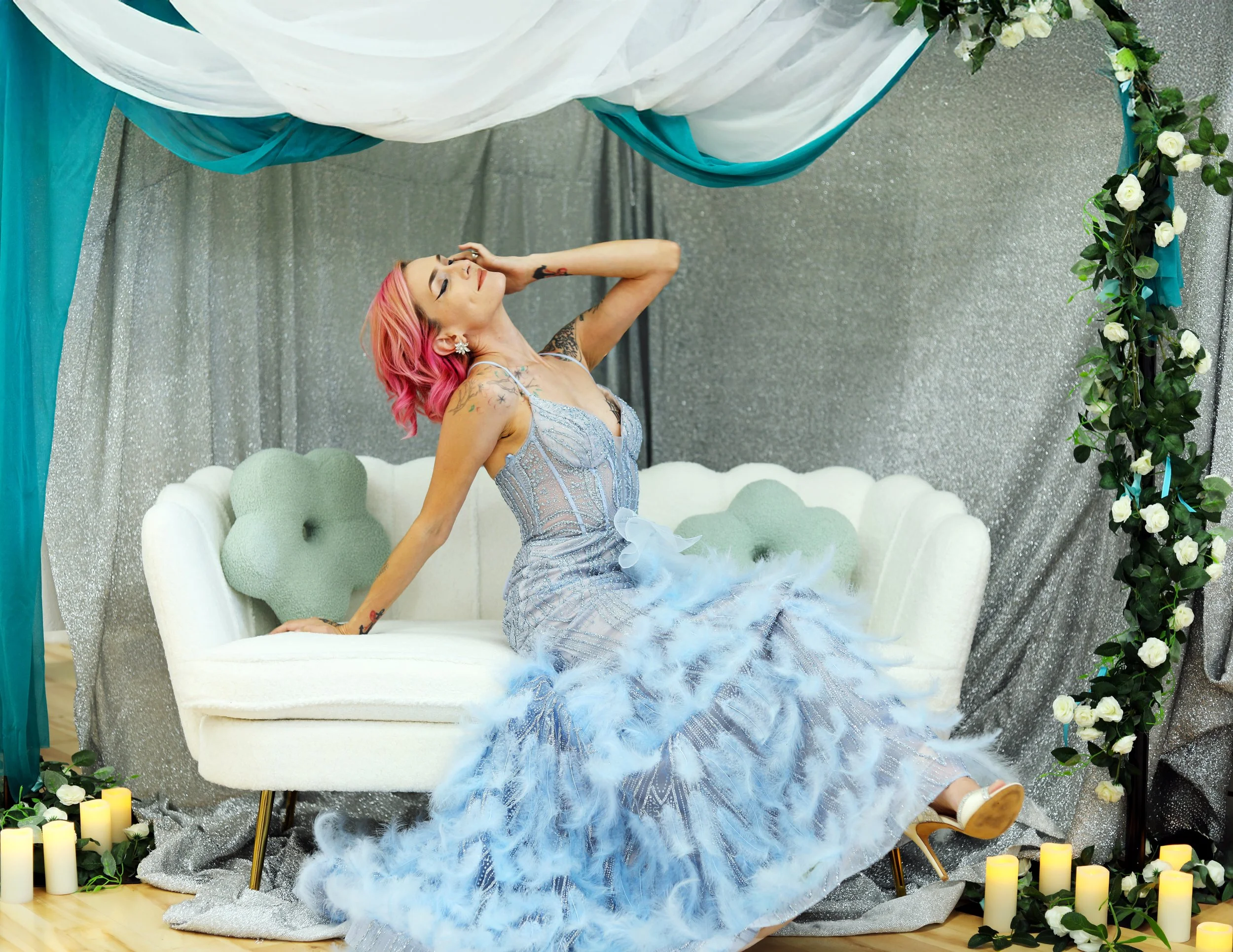 A woman with pink hair and tattoos posing on a white vintage-style sofa with floral and candles decoration.