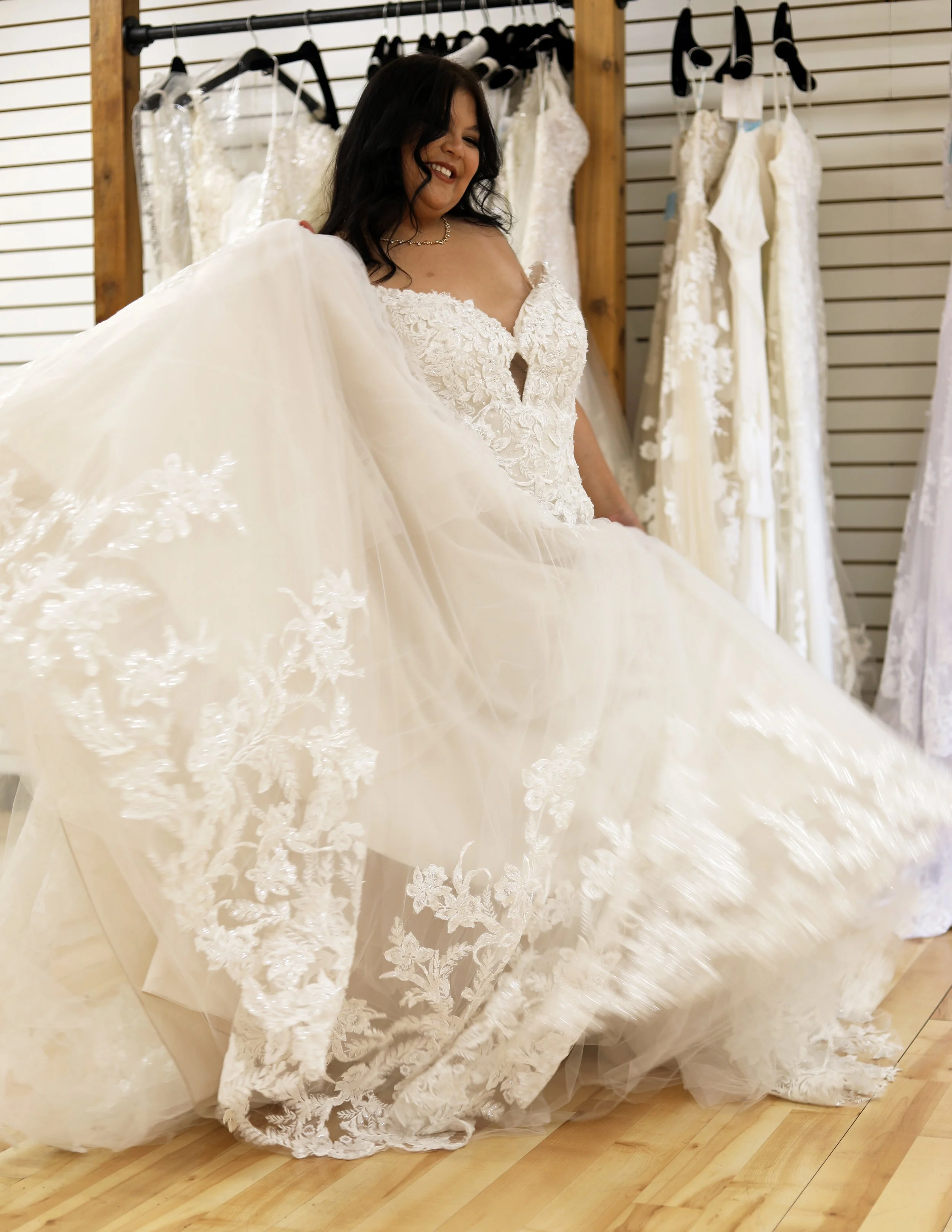 A woman in a bridal boutique trying on a wedding dress with lace details and a full skirt, surrounded by other wedding dresses hanging on racks.