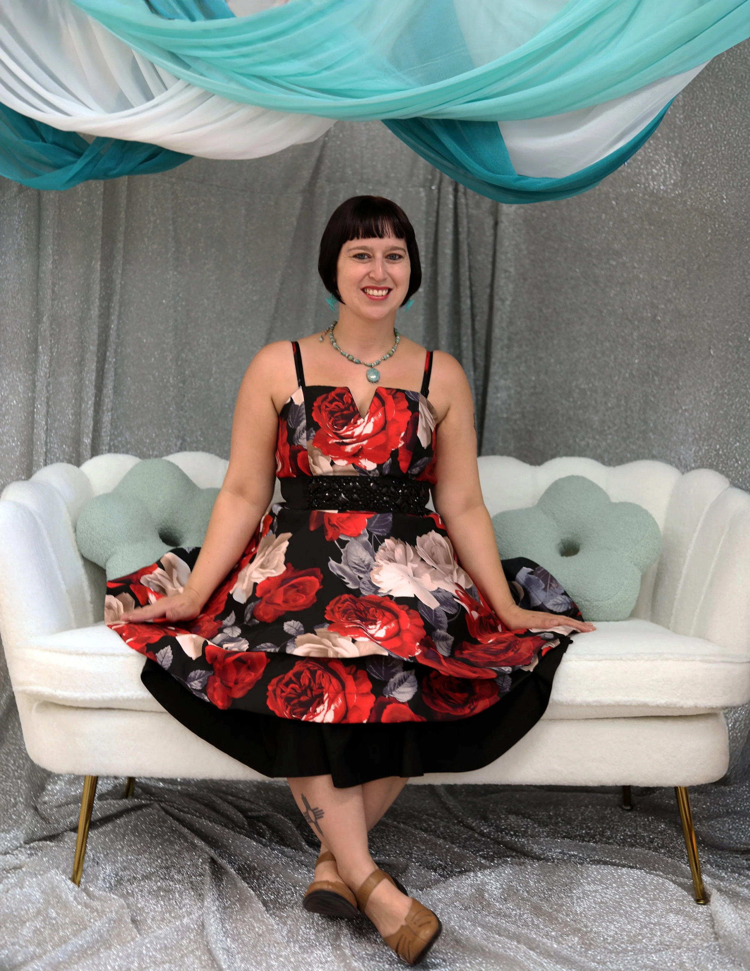 Smiling woman wearing a floral dress sitting on a white vintage-style sofa with a silver glitter backdrop and teal and white draped fabric overhead.