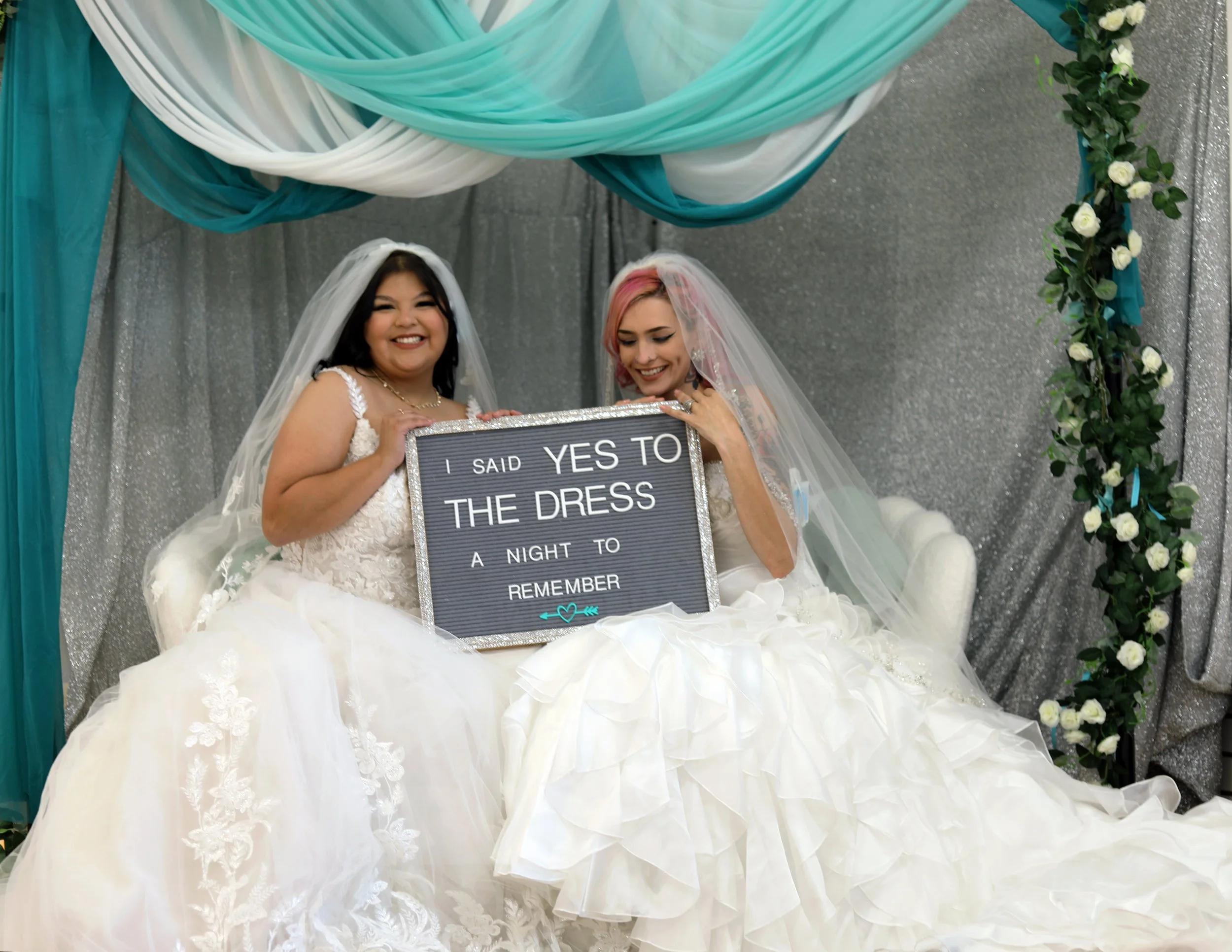 Two women in wedding dresses sitting on a white couch, holding a sign that reads, 'I said YES to the dress, a night to remember.' They are under a decorative canopy with teal, white, and silver drapery and flowers.
