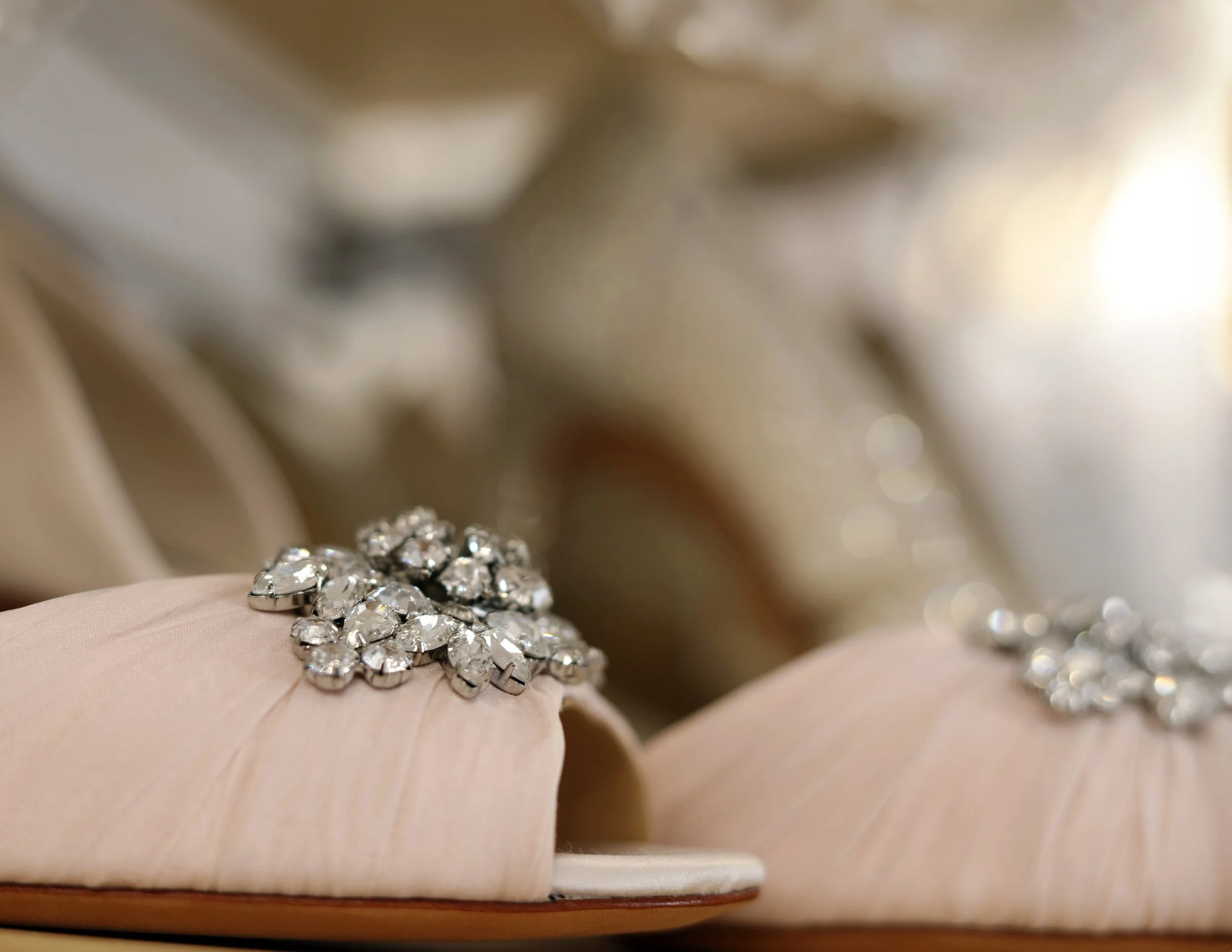 A pair of elegant beige satin shoes adorned with sparkling rhinestone embellishments on the toes, with a blurred background.
