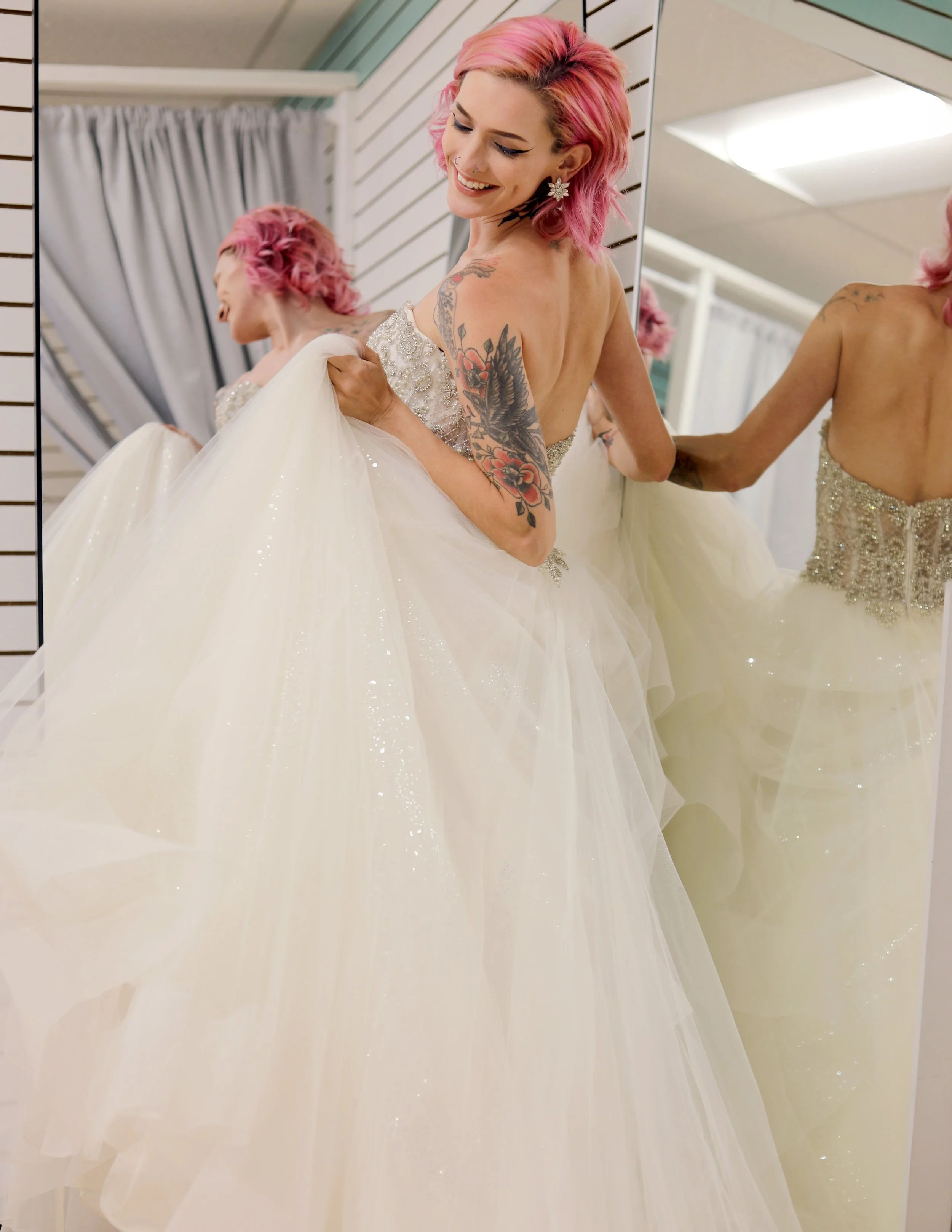 A woman with pink hair putting on a wedding dress in front of a mirror.
