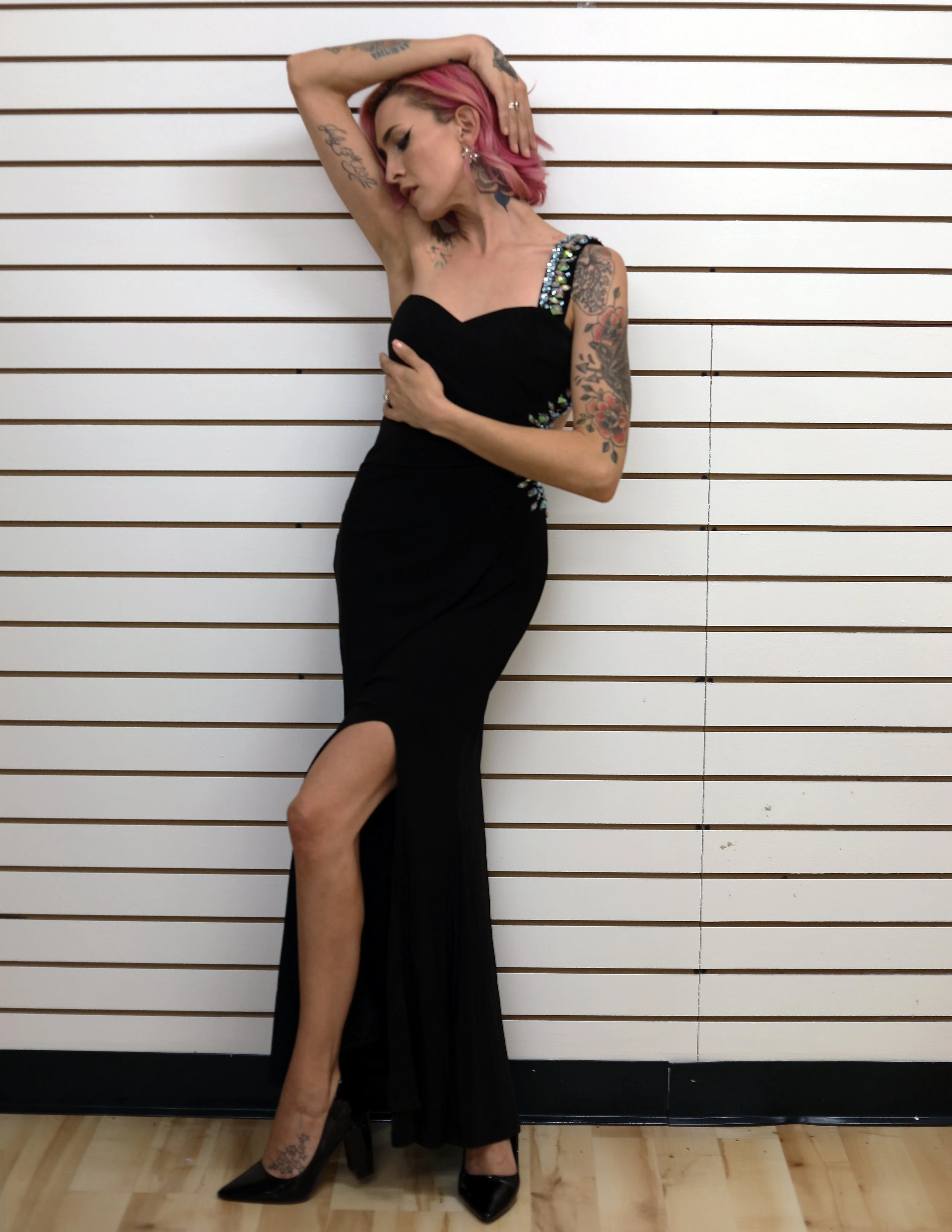 Woman with pink hair wearing a black dress with embellishments, standing against a white slat wall, striking a pose with one arm raised and her head tilted.
