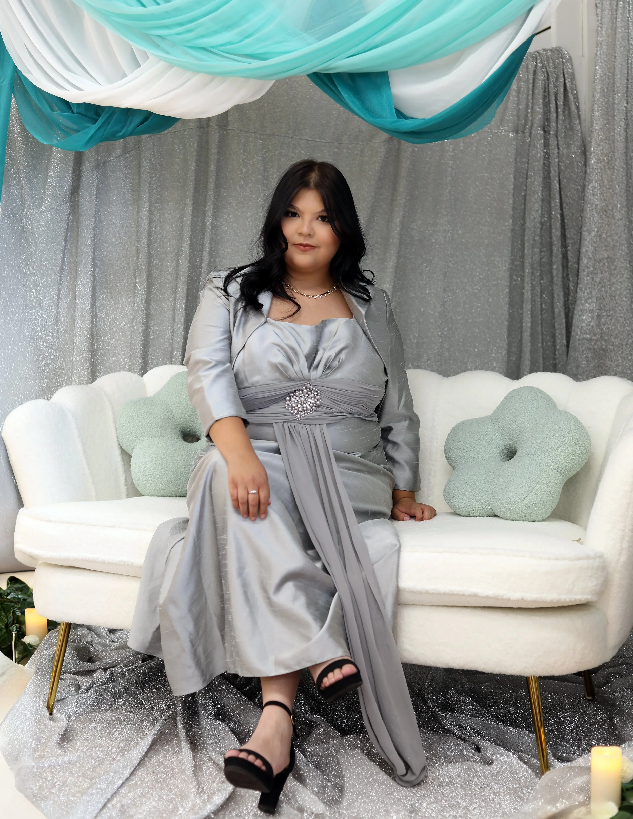 A woman in a silver dress sitting on a white, vintage-style sofa with pastel green and white decorations overhead, silver glittering curtains in the background, and a sparkly silver floor covering.