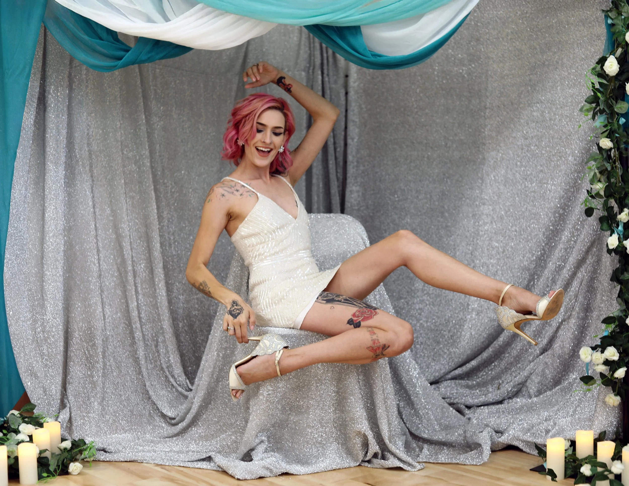 A woman with pink hair posing on a sparkly silver backdrop, surrounded by candles and greenery, wearing a cream-colored dress and high heels.
