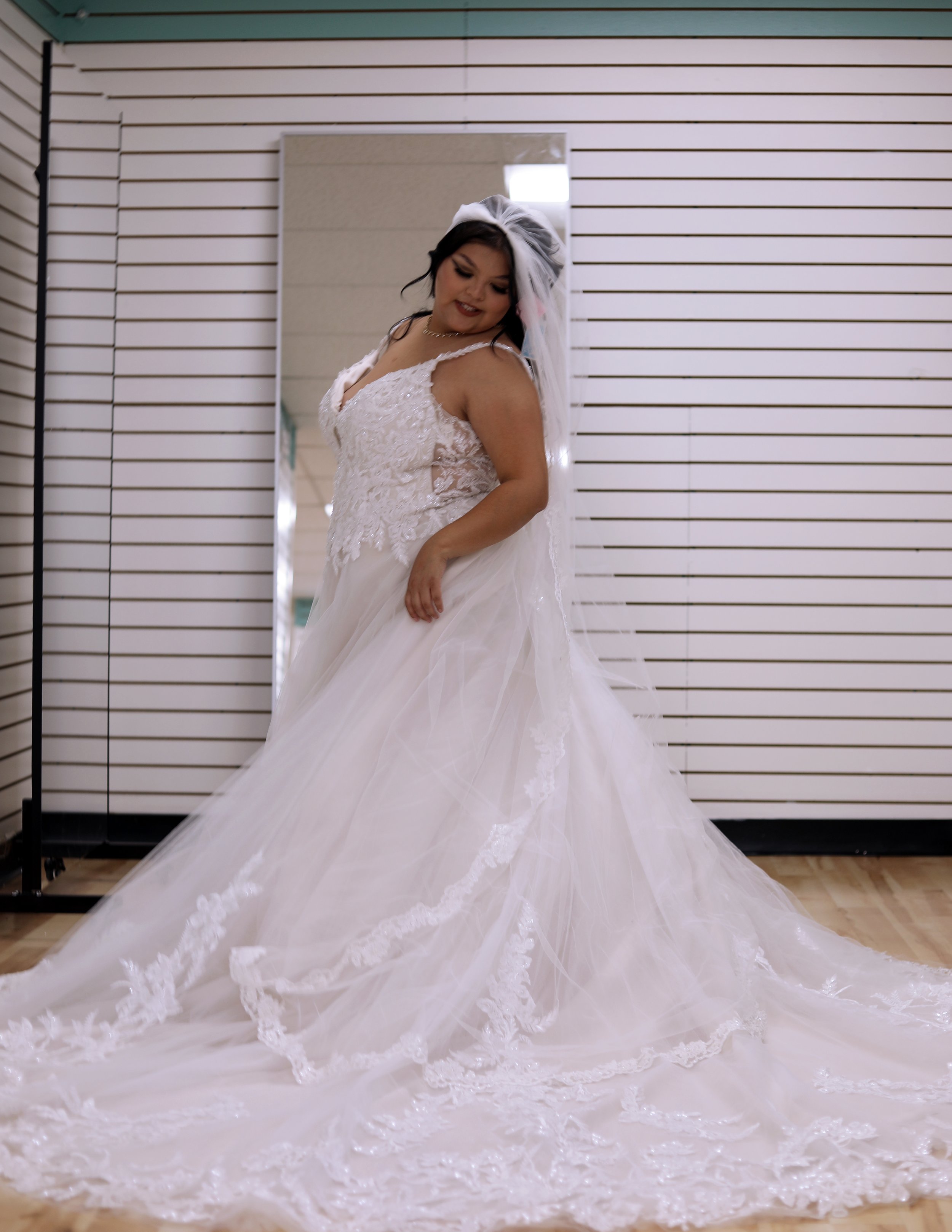 A woman in a white wedding gown with lace details, holding the skirt and smiling, standing in front of a striped wall in a fitting room.