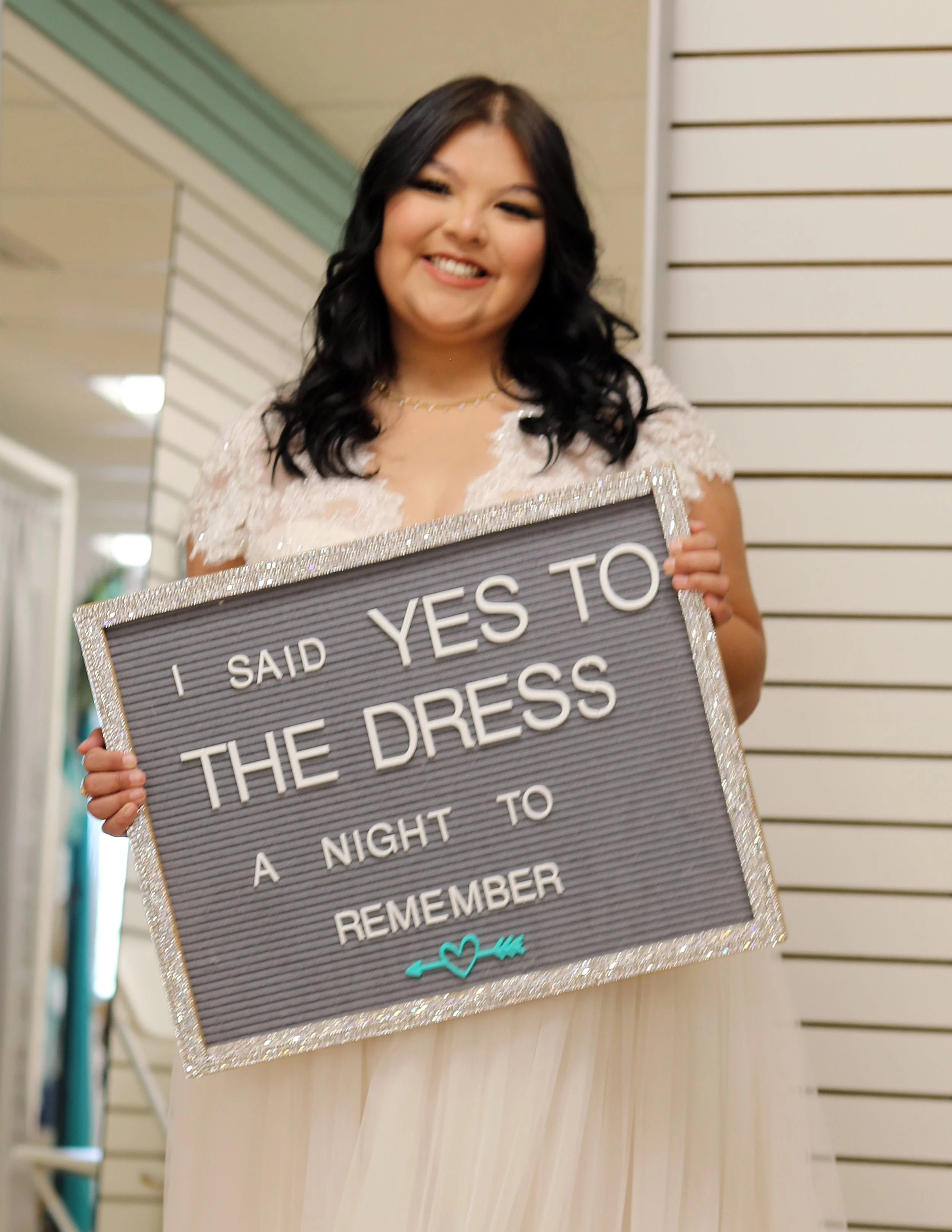 Woman with black hair smiling and holding a sign that says, 'I said yes to the dress, a night to remember,' in an indoor setting.