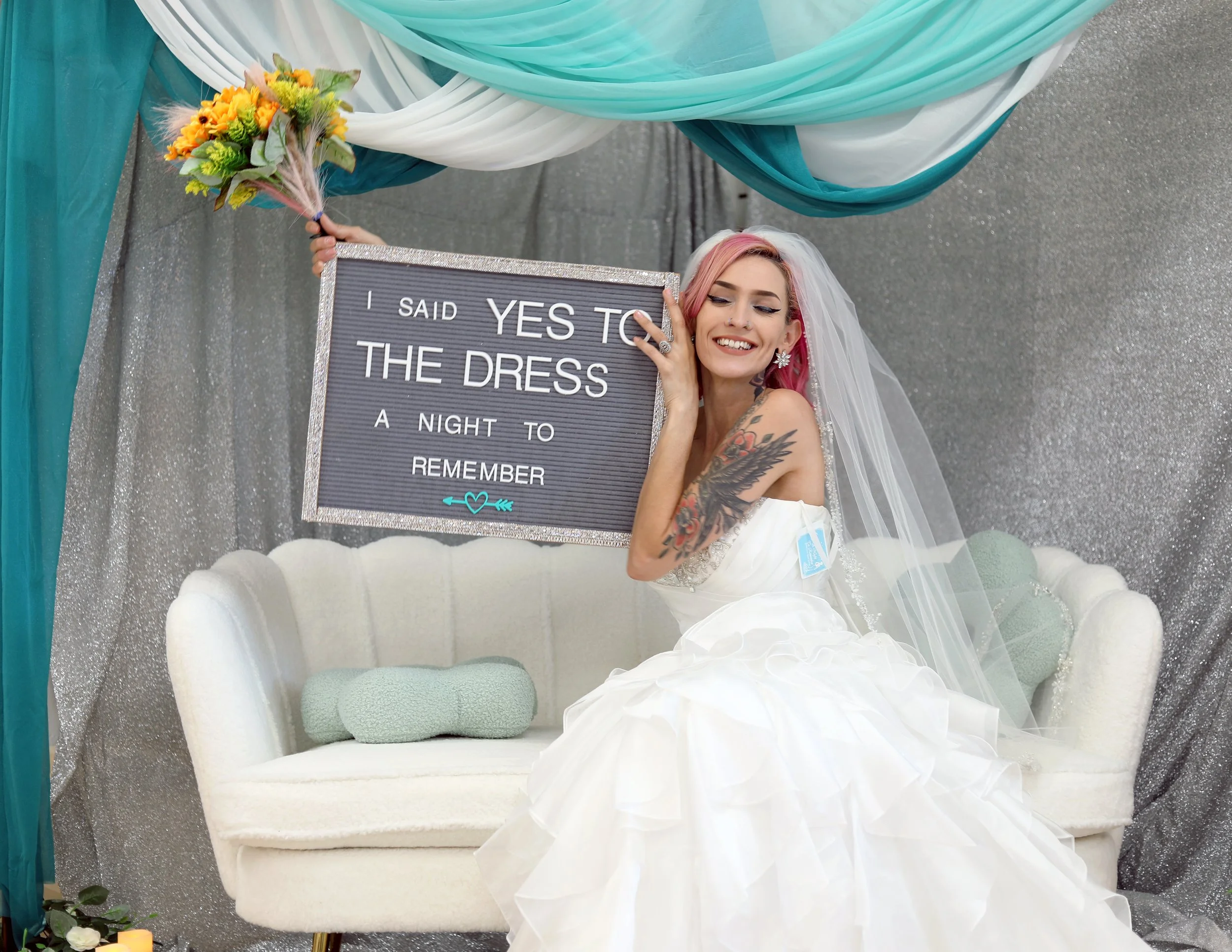 A woman in a wedding dress with pink hair and tattoos, sitting on a white vintage sofa, holding a sign that says "I said YES to the dress, a night to remember," and smiling with her eyes closed. She is wearing a veil and earrings, with a colorful bou