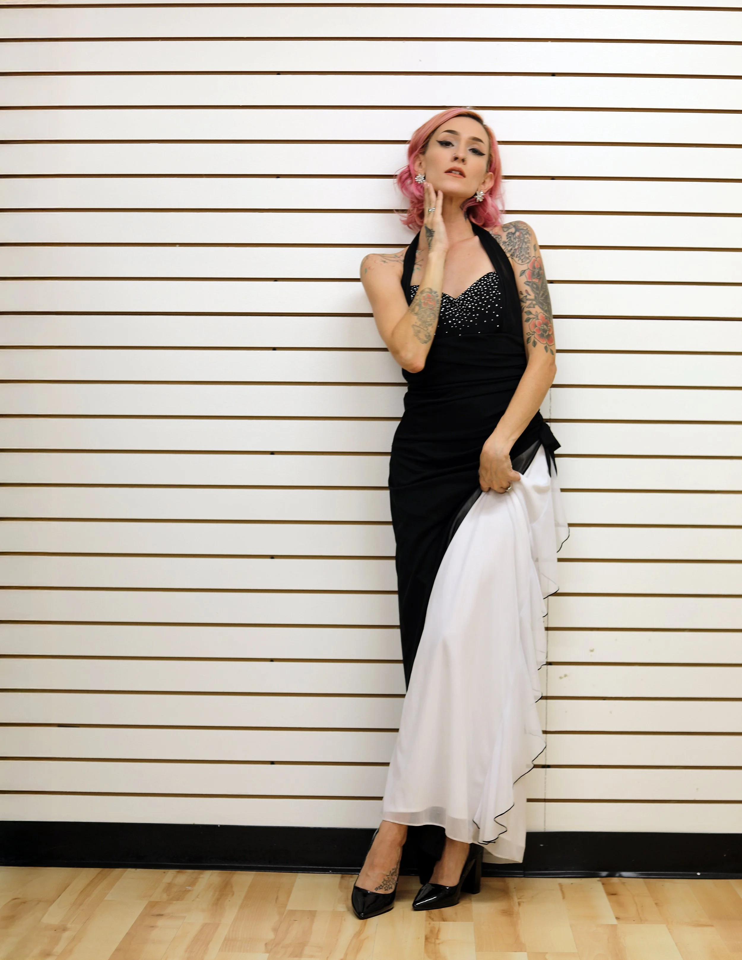 A woman with pink hair and tattoos standing against a striped wall in a black and white dress with black high heels.