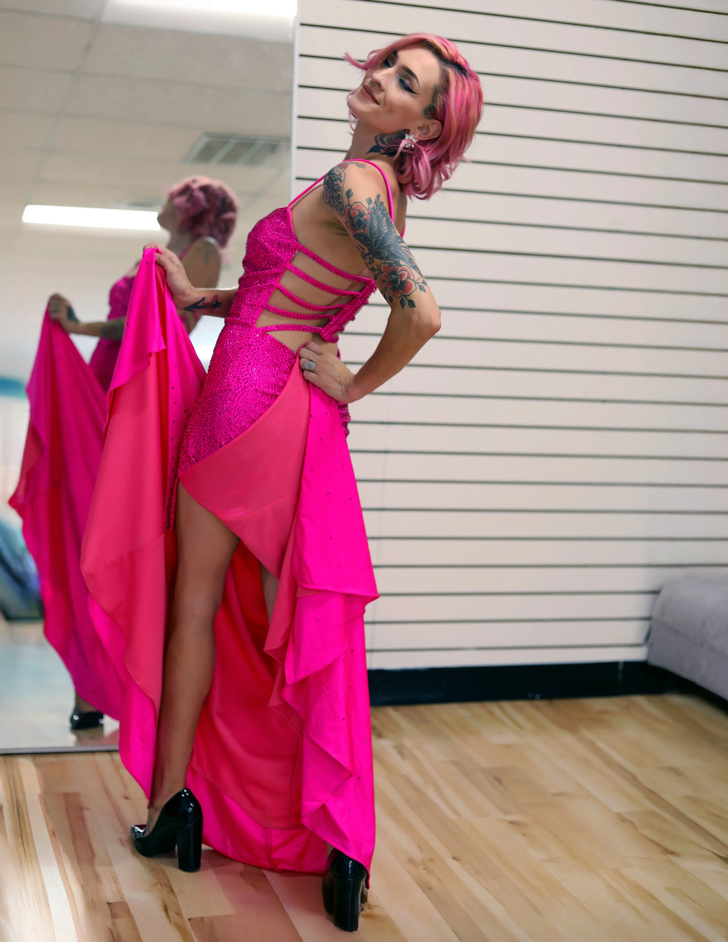 A woman with pink hair and tattoos is wearing a bright pink, sparkly dress with cutouts on the side. She is standing in front of a mirror, holding part of her dress, and smiling with her eyes closed.