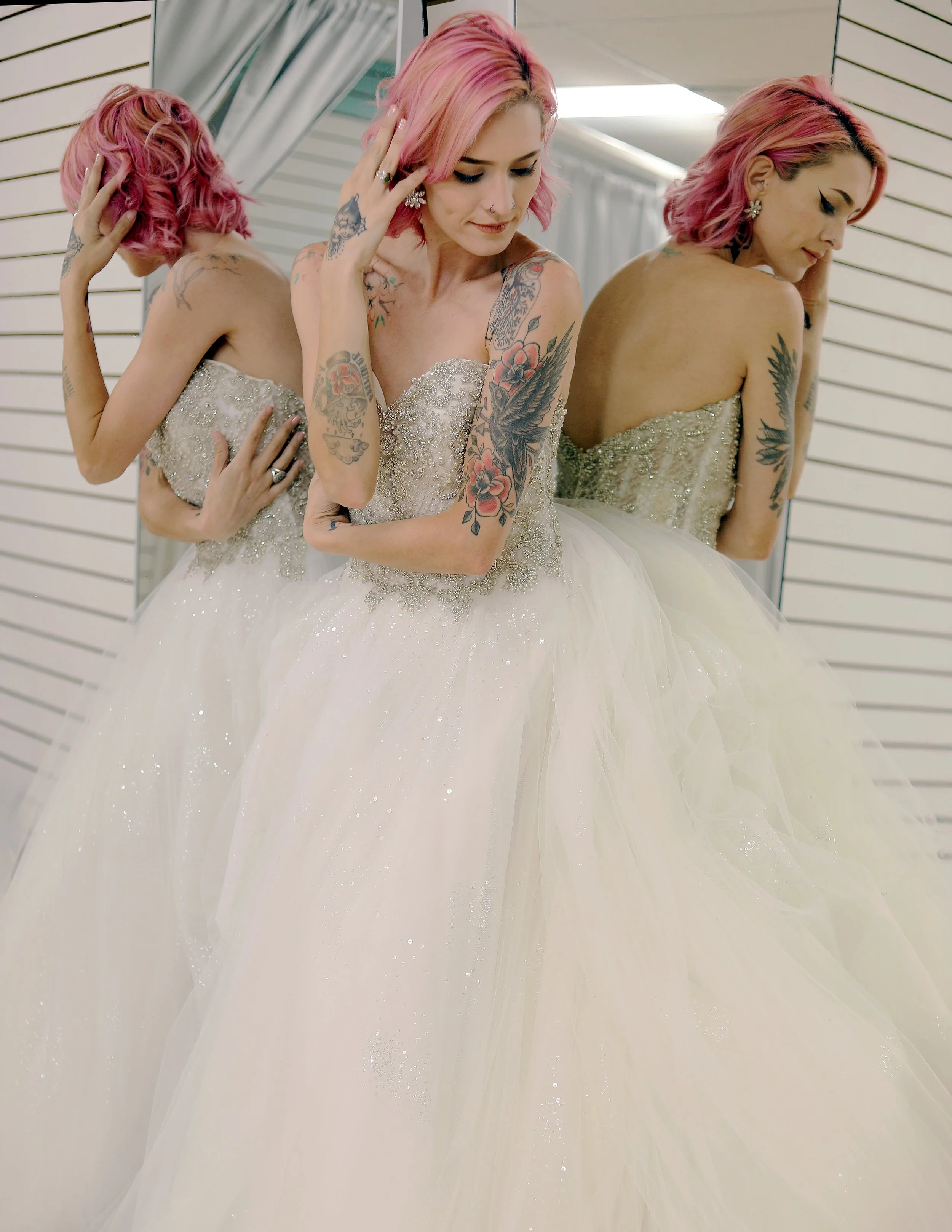 A woman with pink hair trying on a wedding dress, standing in front of a mirror with reflections of herself.