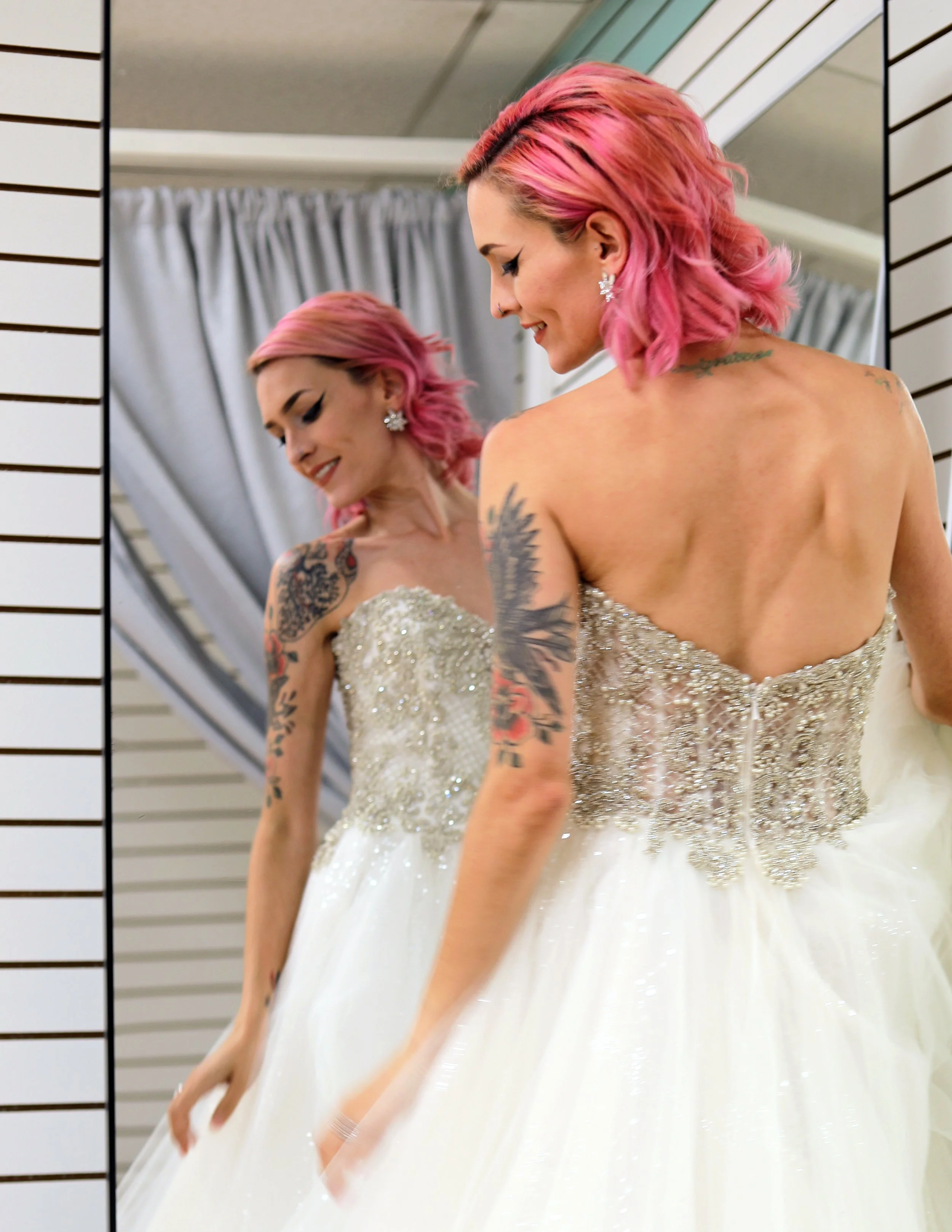 A woman with pink hair and tattoos trying on a white wedding dress in front of a mirror.