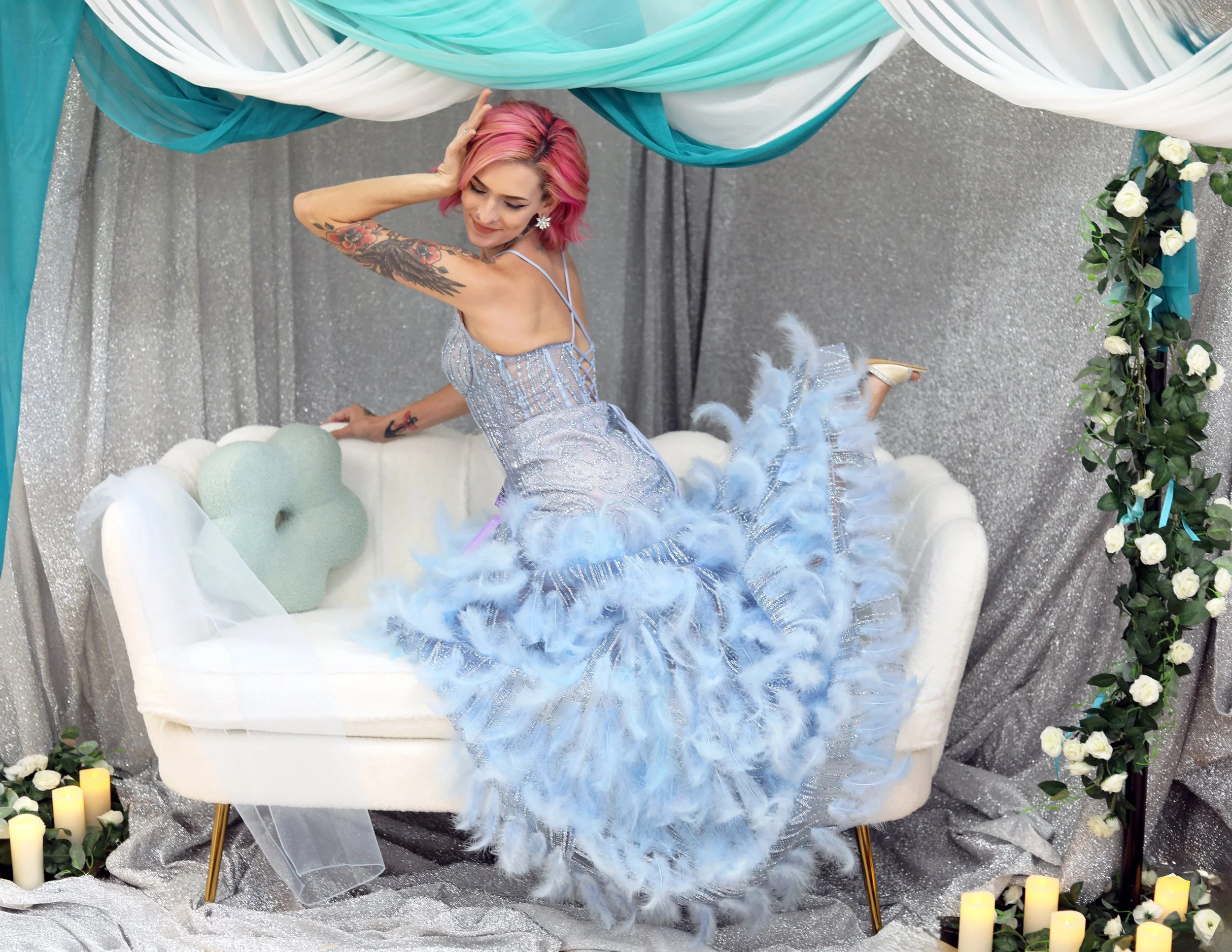 Woman with pink hair and tattoos on her arm, wearing a shimmering gown with feathers, sitting on a white tufted sofa decorated with candles and flowers, with teal and silver fabric drapes hanging overhead and a silver glitter backdrop.