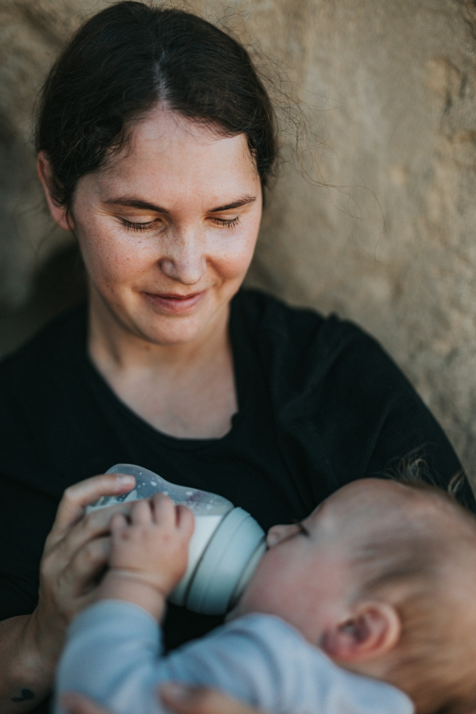 A woman with dark hair feeding a baby with a bottle, both laying on a soft surface near a stone wall.