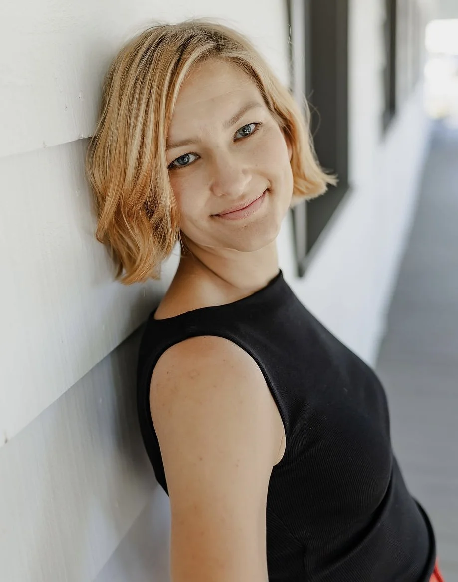 A young woman with short, wavy blonde hair and blue eyes smiling while leaning against a white wooden wall near a window.
