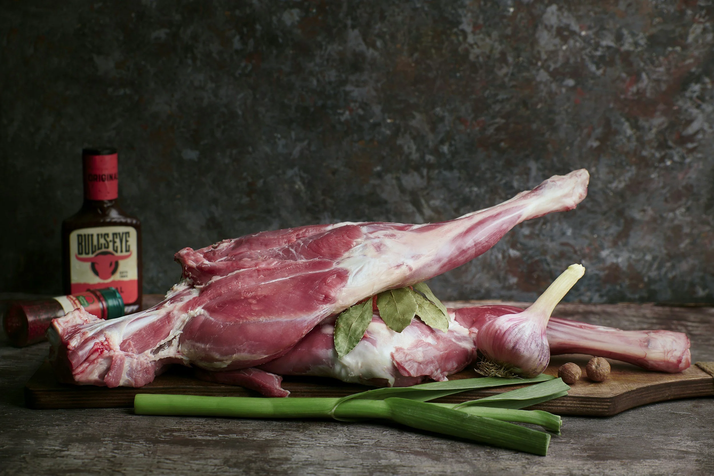 Raw lamb meat on a wooden cutting board with garlic, green onions, bay leaves, and spices, in front of a dark textured background.