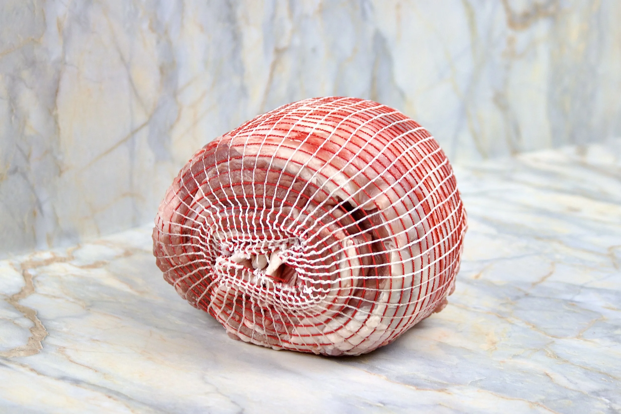 A raw piece of meat, likely beef, wrapped in a netting, resting on a marble surface with a marble background.