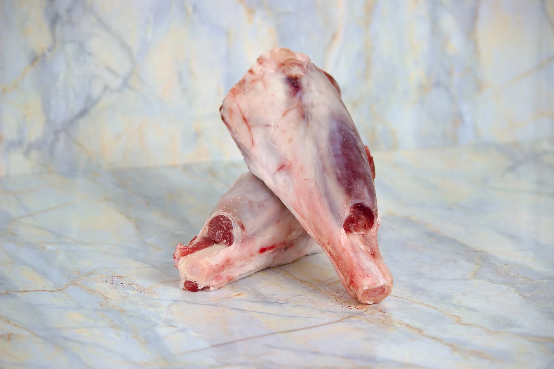 Two raw lamb shanks placed on a marble surface with a marble background.