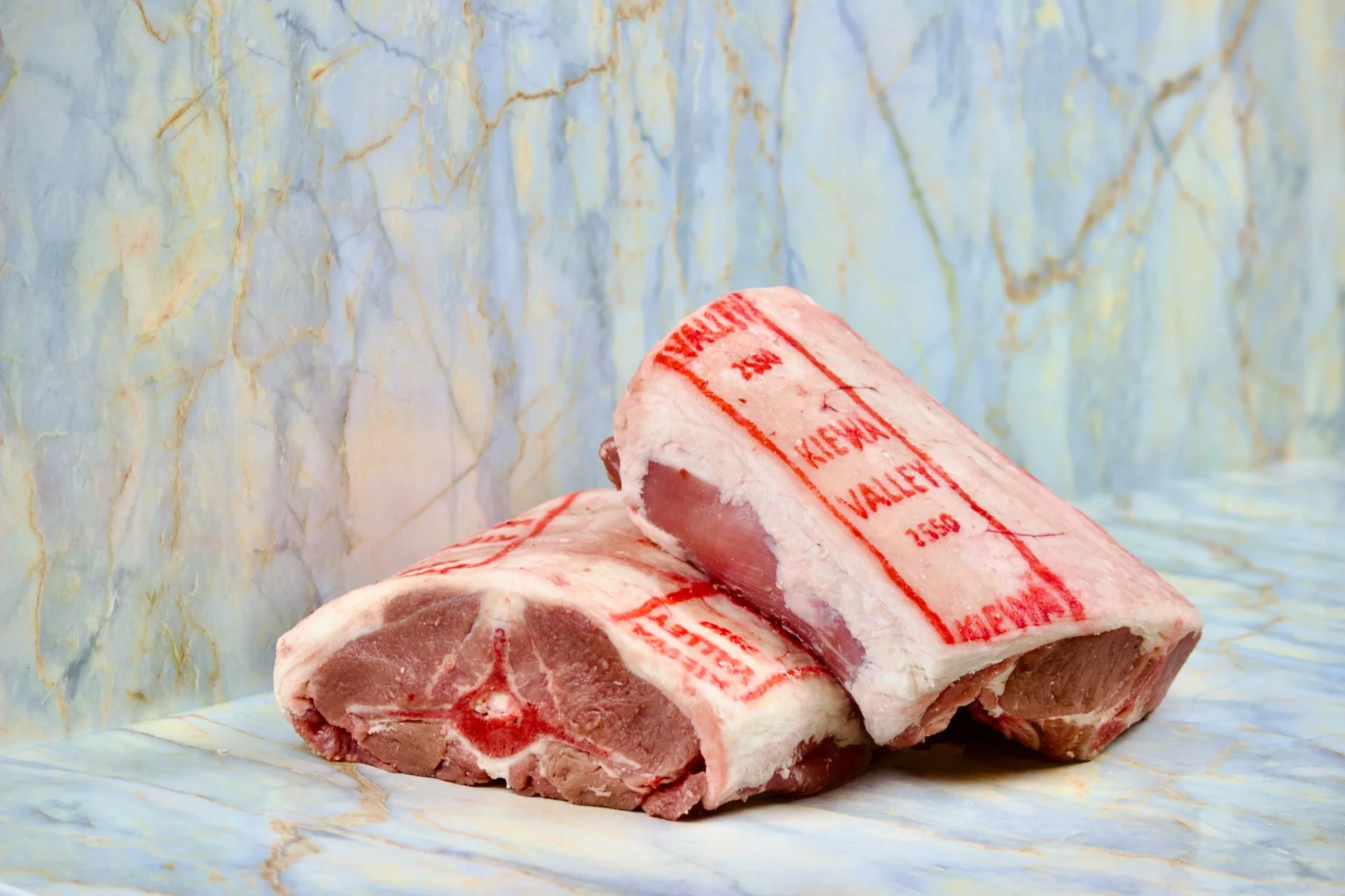 Two raw bone-in steaks with red 'inspection' stamps on the packaging, resting on a marble surface with a marble background.