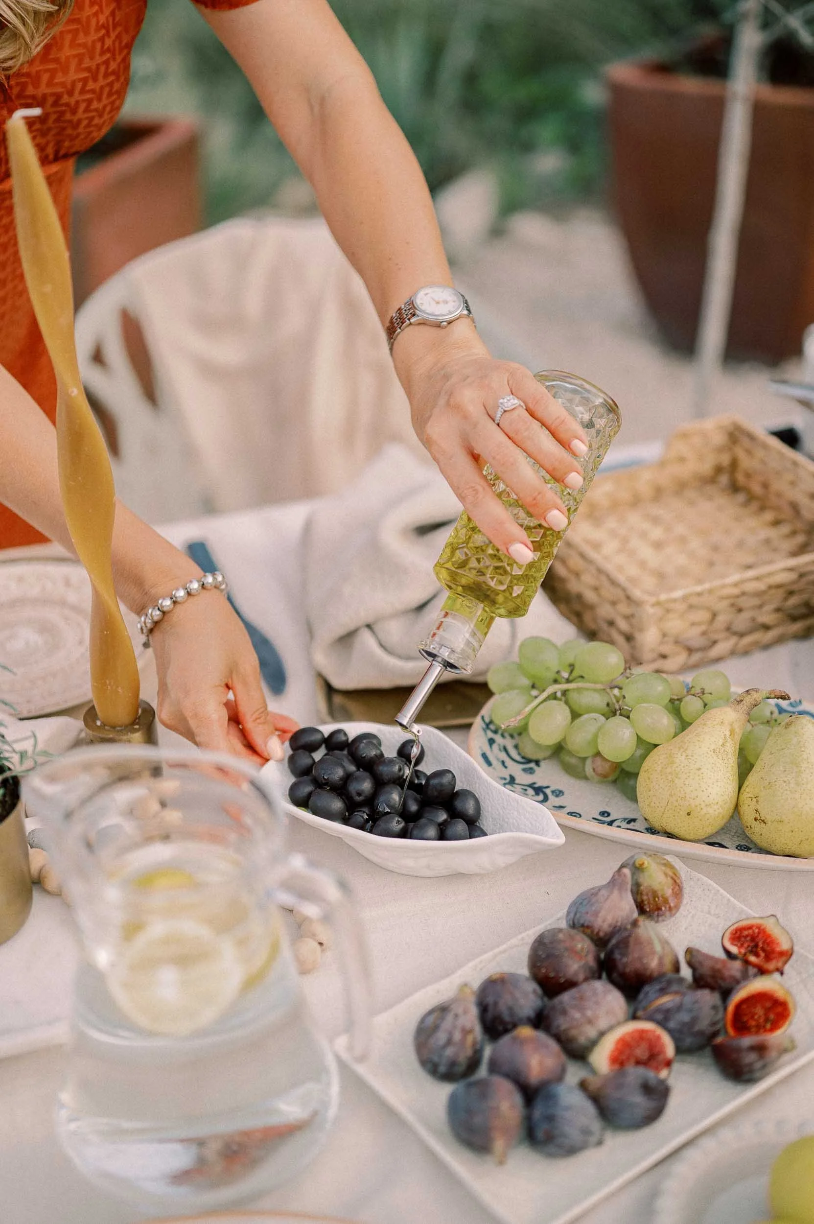 Person pouring olive oil over black grapes at a table with various fruits including pears, figs, and grapes, with decorative items in the background.