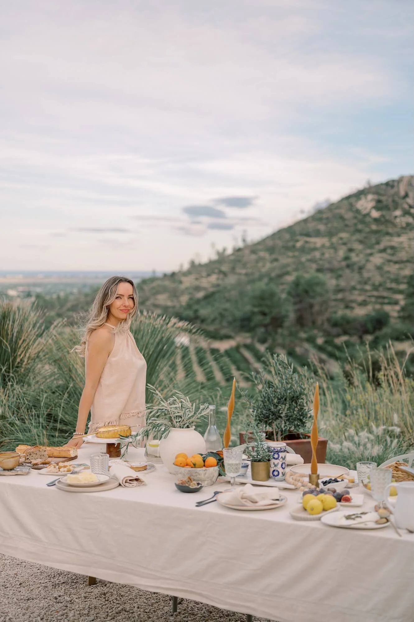 Denisa standing next to an outdoor dining table set with food and candles, with a scenic hillside and cloudy sky in the background.