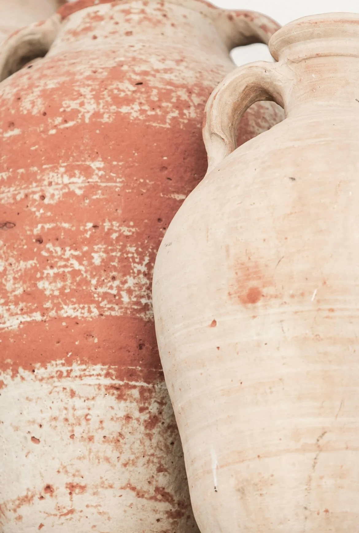 Close-up of two ancient clay pottery jars, one reddish-brown with weathered surface, the other cream-colored with a smooth surface, against a blurry background.