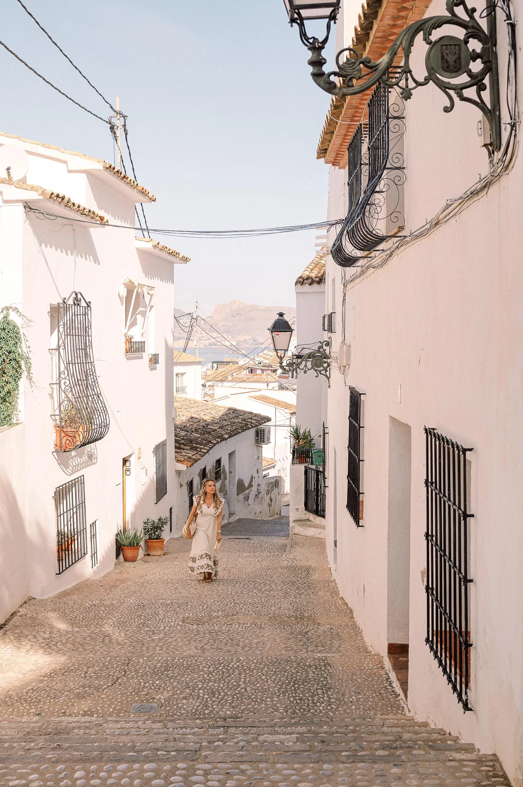 A woman walking up a cobblestone street in a Mediterranean town with white buildings, wrought iron balconies, and potted plants.