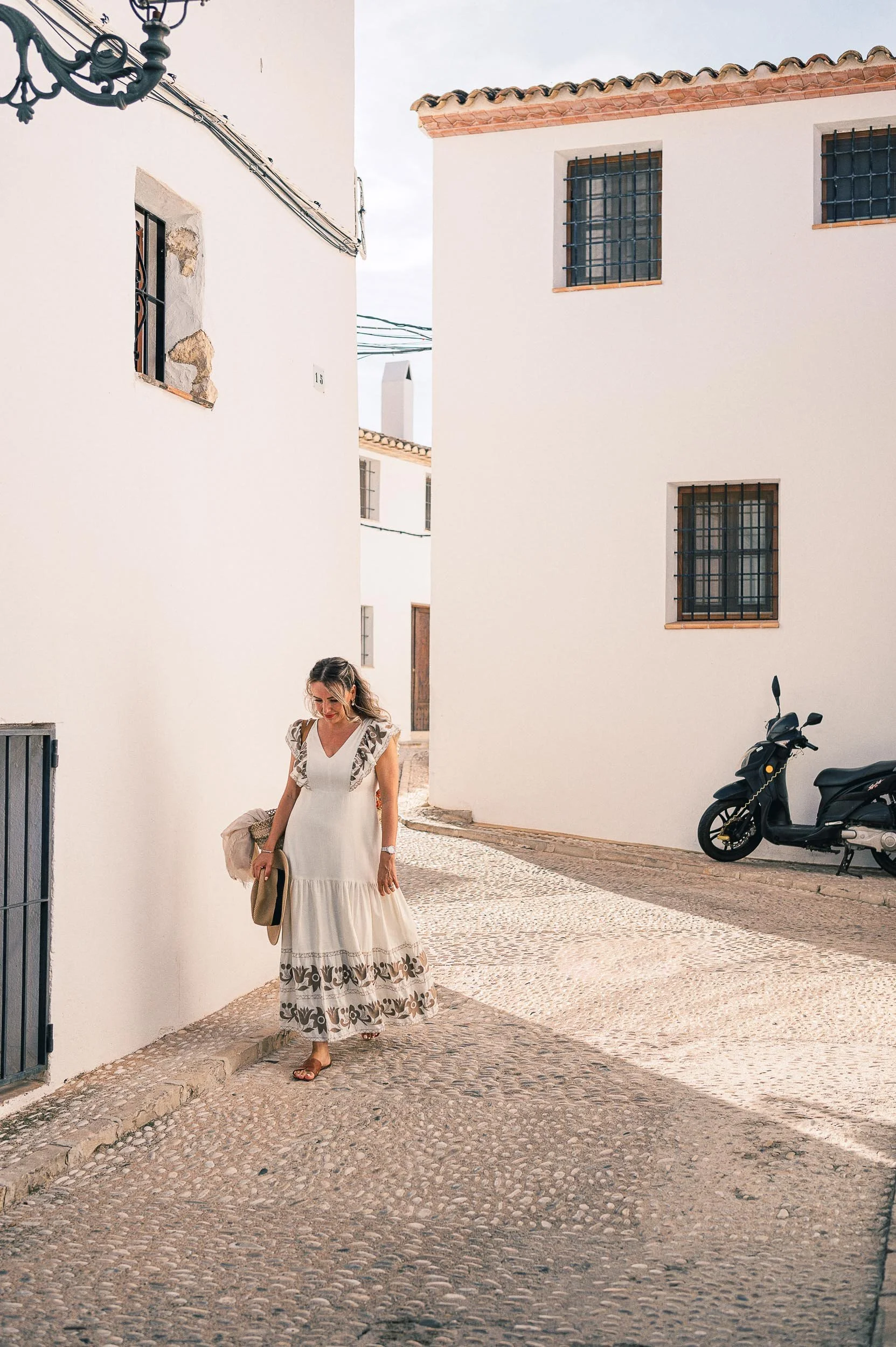 Denisa in a white dress with black patterns on the hem and, holding a hat and a bag, walking in a narrow cobblestone street with white buildings and barred windows, with motorcycle parked along the wall.