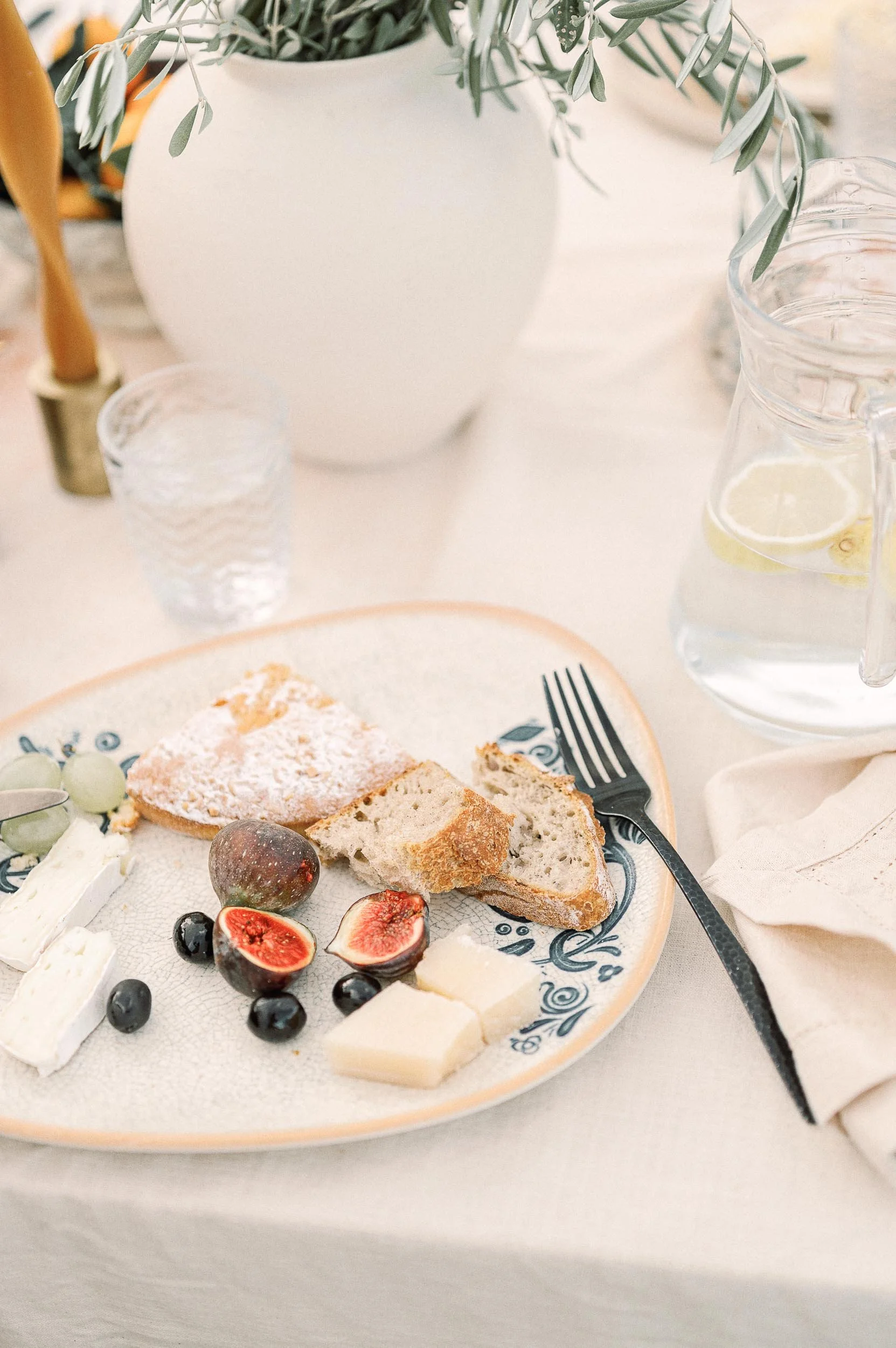 A breakfast or brunch table featuring a plate with slices of bread, figs, grapes, cheese, and a powdered pastry, along with a glass of water with lemon slices, a pitcher of water, a white vase with greenery, and a fork on a napkin.