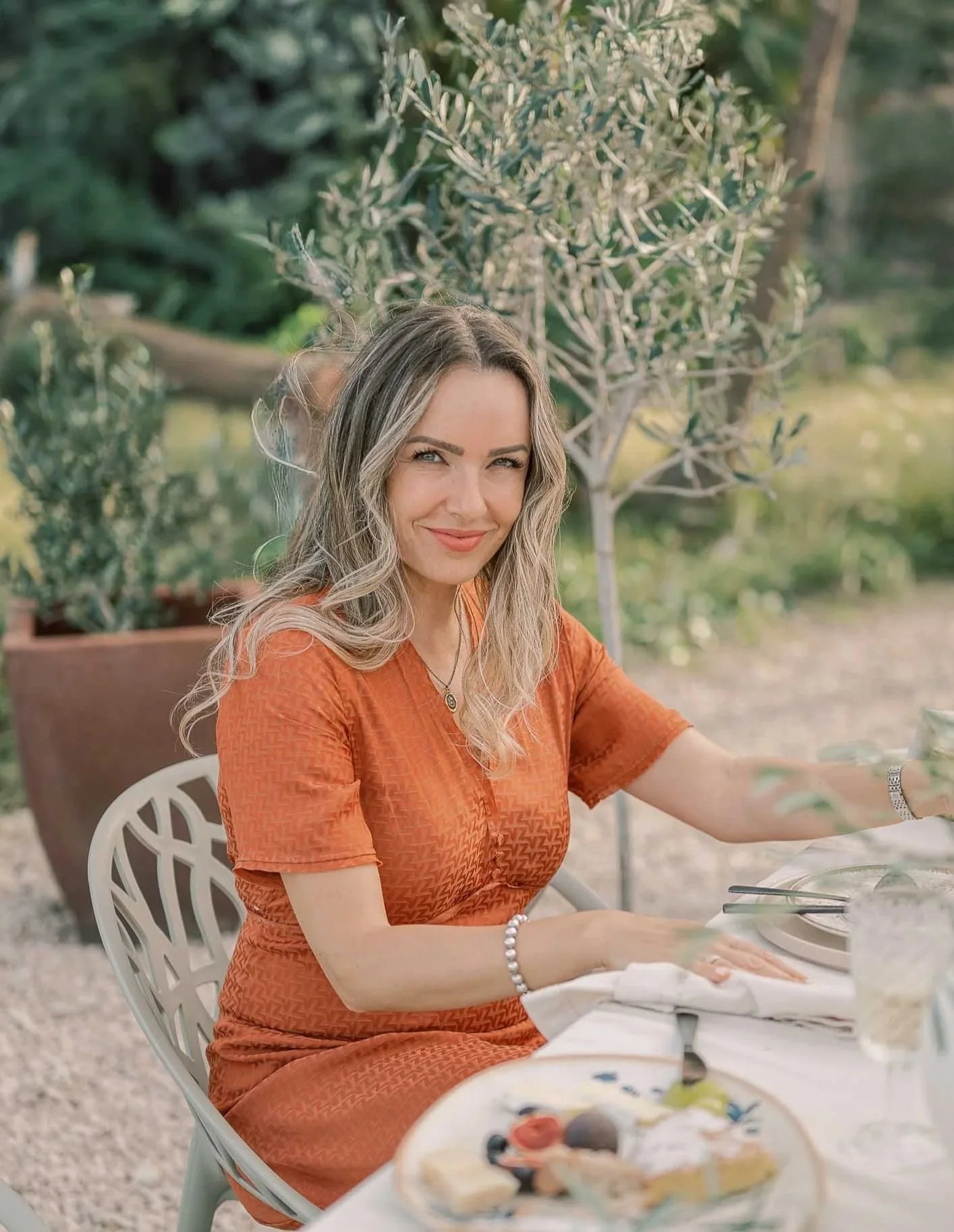 Denisa with wavy blonde hair wearing an orange dress sitting at an outdoor table set for a meal, smiling at the camera, with a garden and potted plants in the background.
